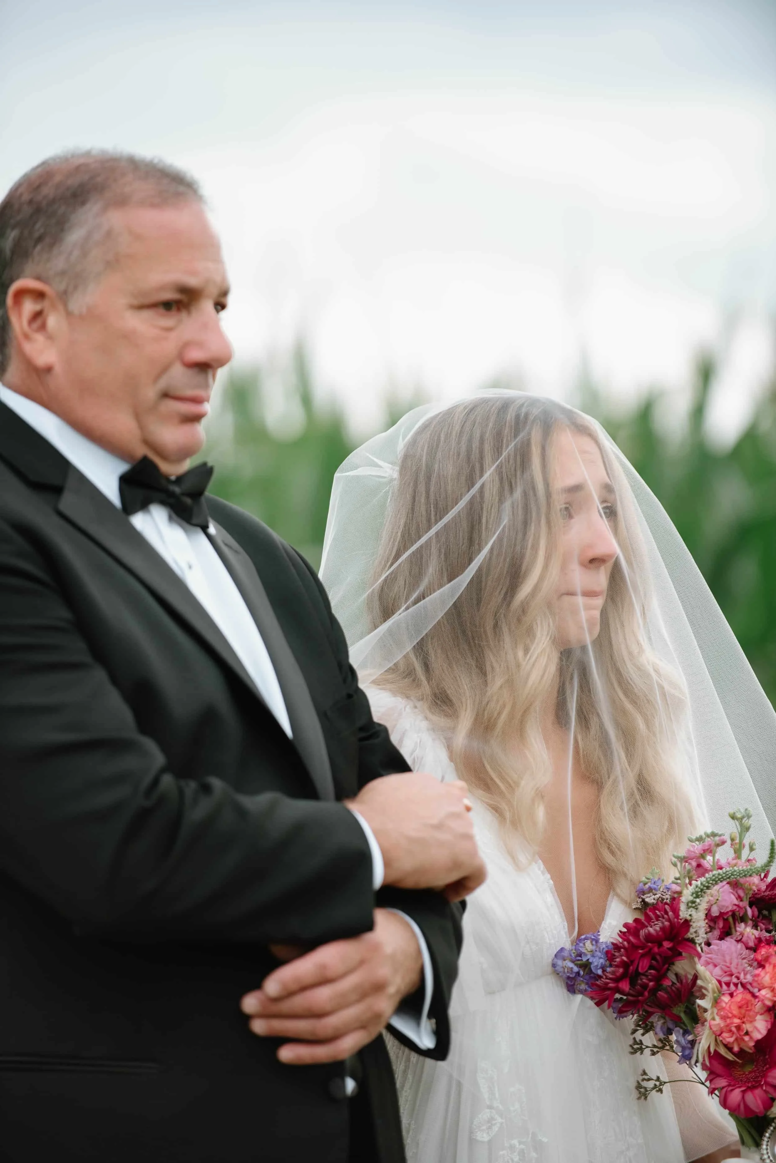 A groom in a black tuxedo with a bowtie standing next to a bride in a white wedding dress holding a bouquet, both with serious expressions. The bride is wearing a veil, and they are outdoors with a green field in the background.
