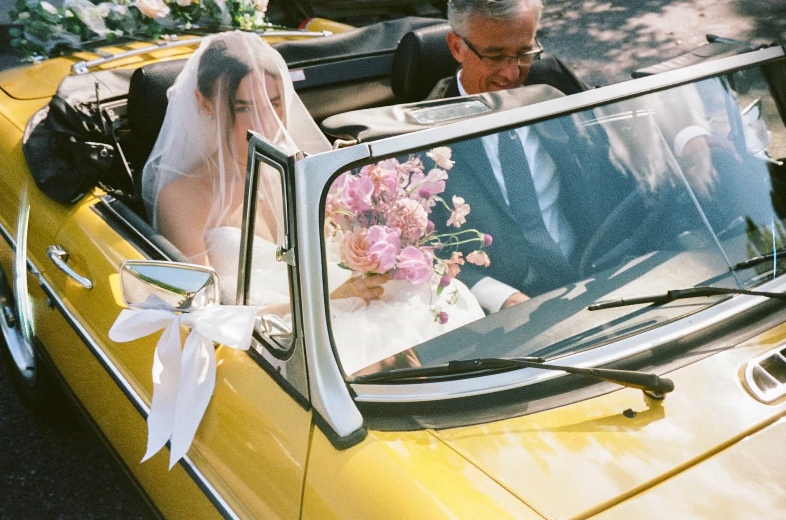 A bride with a veil and white dress sitting in a yellow convertible car, holding a bouquet of pink flowers. She is beside her groom, who is wearing glasses and a suit. The car has a white ribbon tied to the side mirror.
