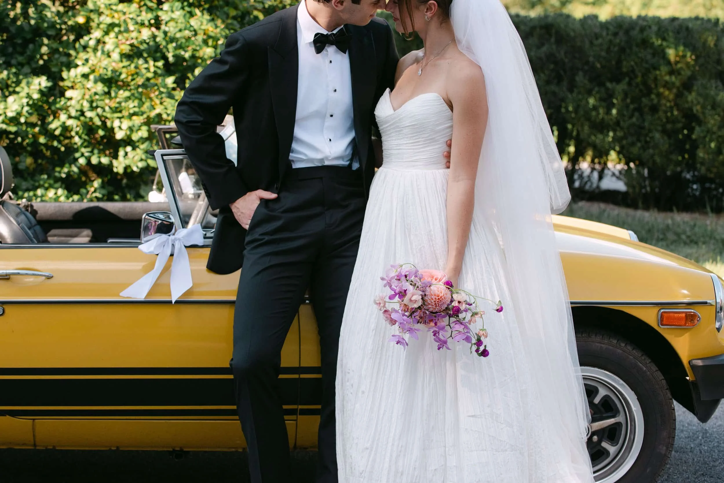 Bride and groom standing close together in front of a yellow vintage car decorated with a white ribbon, with greenery in the background.