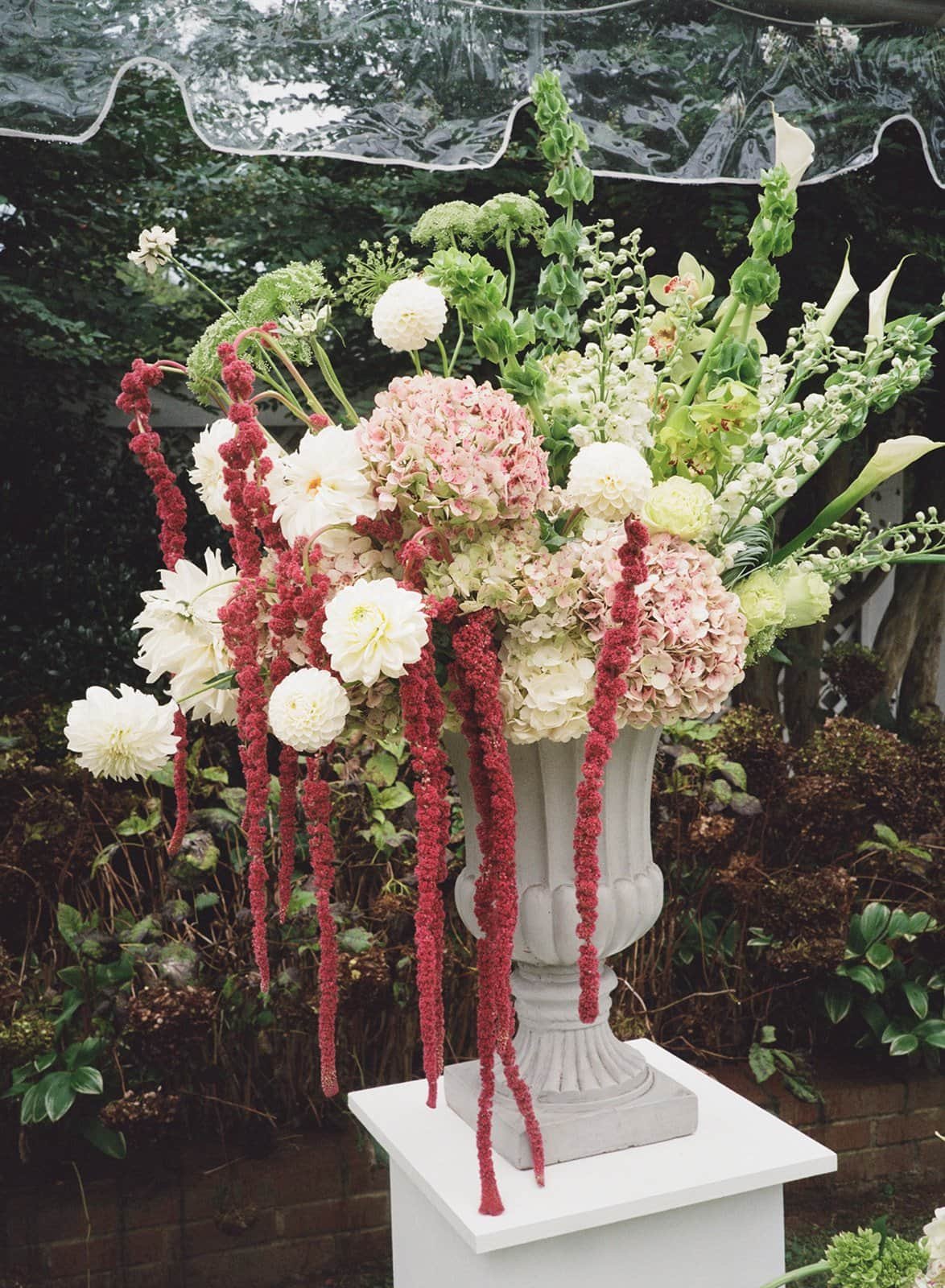 A large floral arrangement in a white urn on a pedestal, featuring pink and white flowers with long, hanging red amaranthus, set outdoors against greenery and a brick wall at The Clifton Inn in Charlottesville Virginia