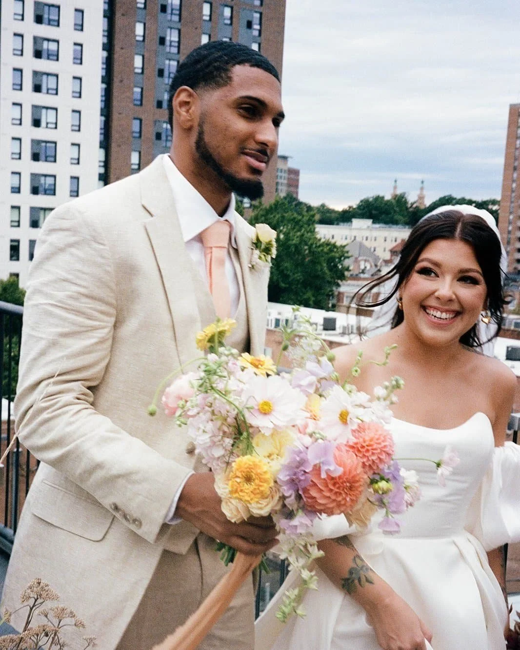 A man and woman in wedding attire smiling on a rooftop with city buildings in the background. The man is holding a colorful bouquet of flowers.