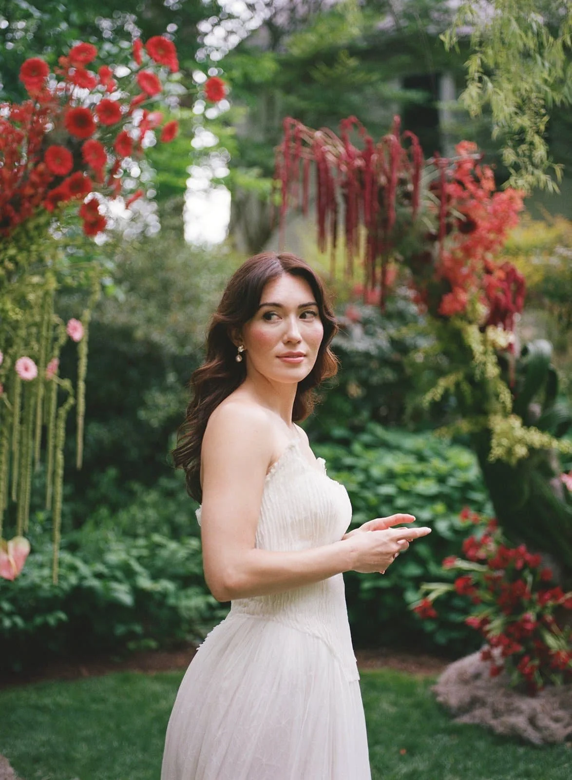 A woman in an elegant off-shoulder white dress standing in a lush garden with colorful flowers and greenery at The Clifton Inn in Charlottesville Virginia