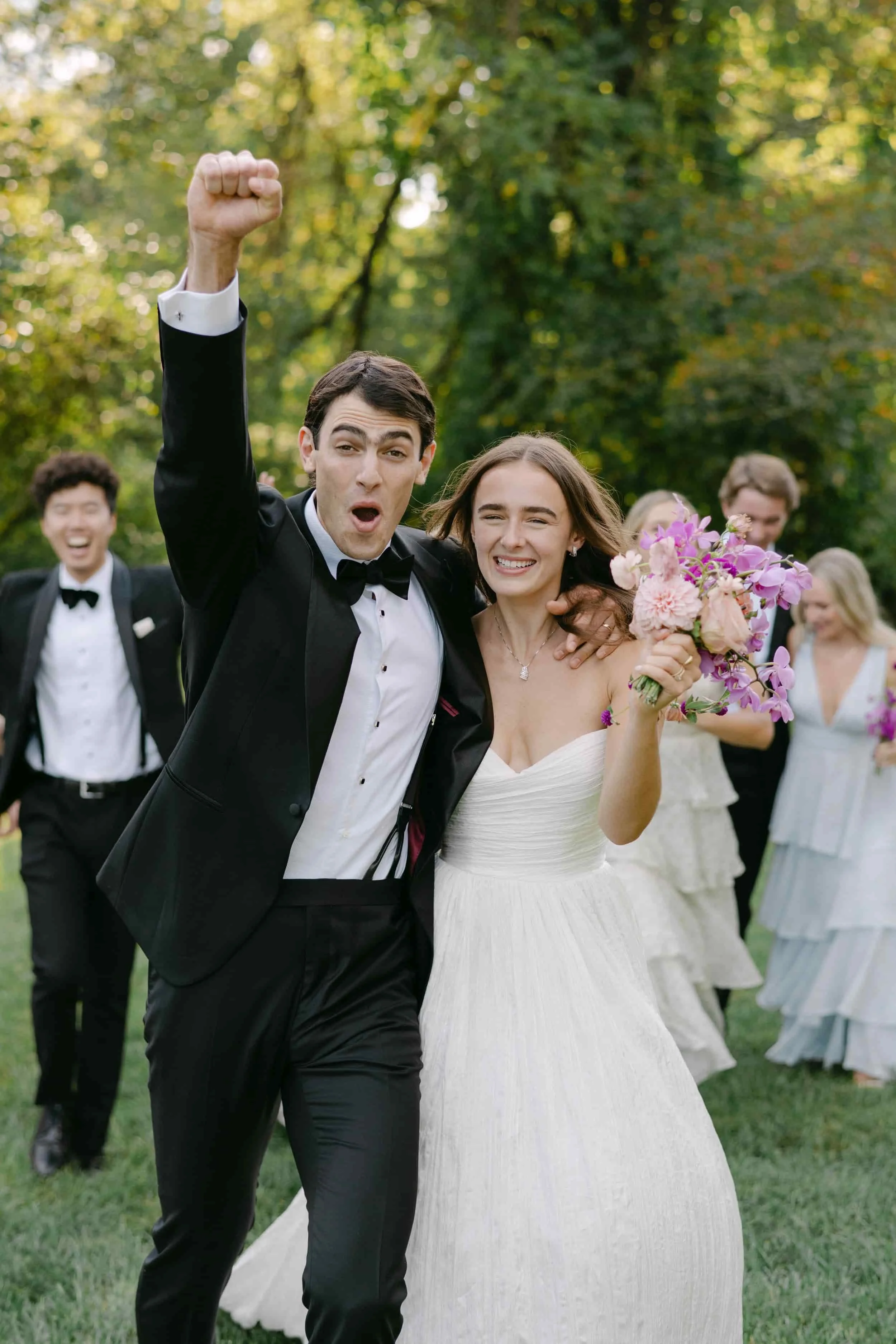 A newly married couple celebrating outdoors in a park, with the groom raising his arm in victory, and friends in the background.