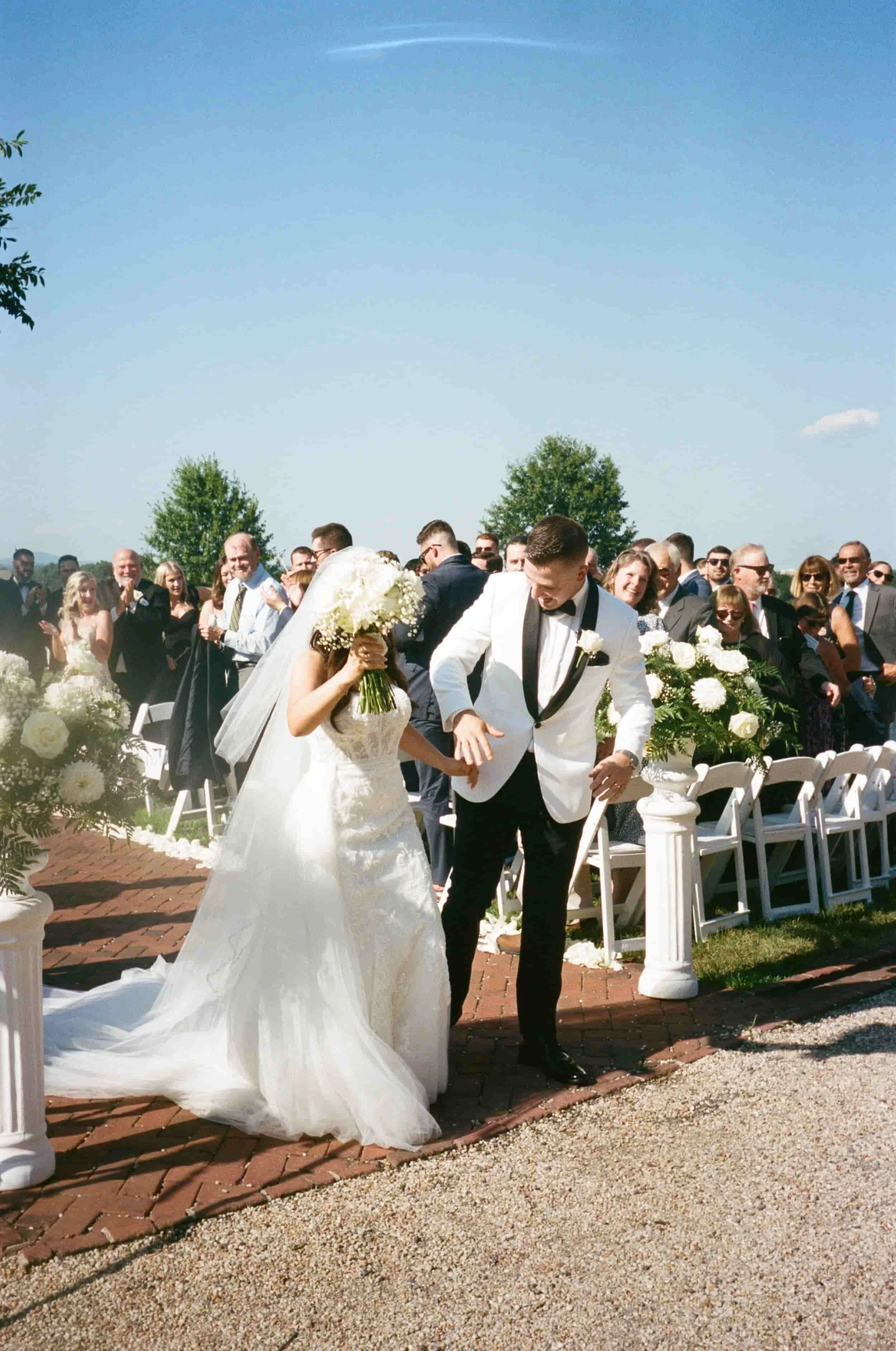 A bride and groom walking down the aisle at an outdoor wedding ceremony with guests clapping and smiling in the background. The bride is covering her face with a bouquet of flowers, and both are dressed in formal wedding attire. The scene is sunny wi