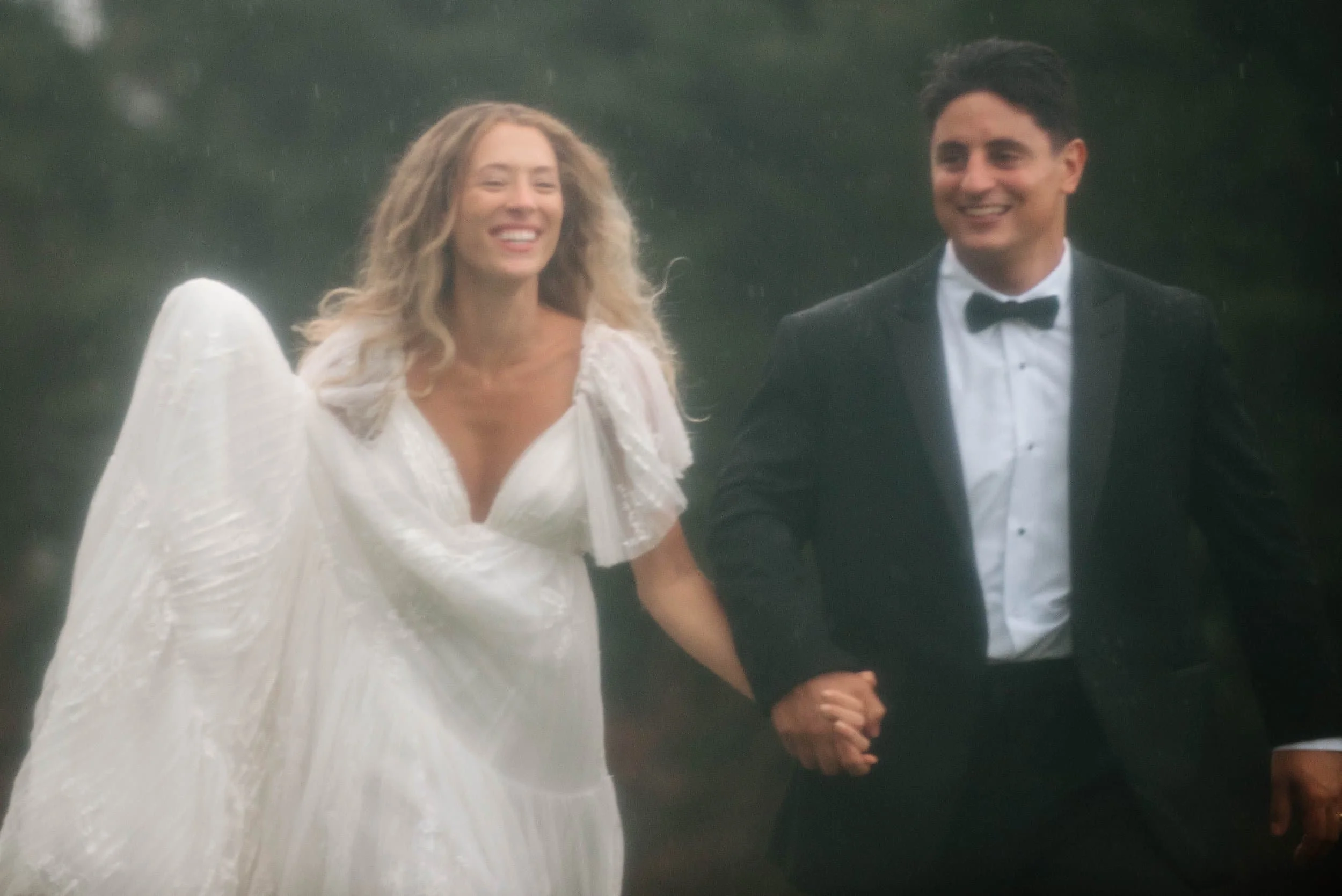 A smiling woman in a wedding dress holding hands and walking with a man in a black tuxedo and bow tie, outdoors in a natural setting.