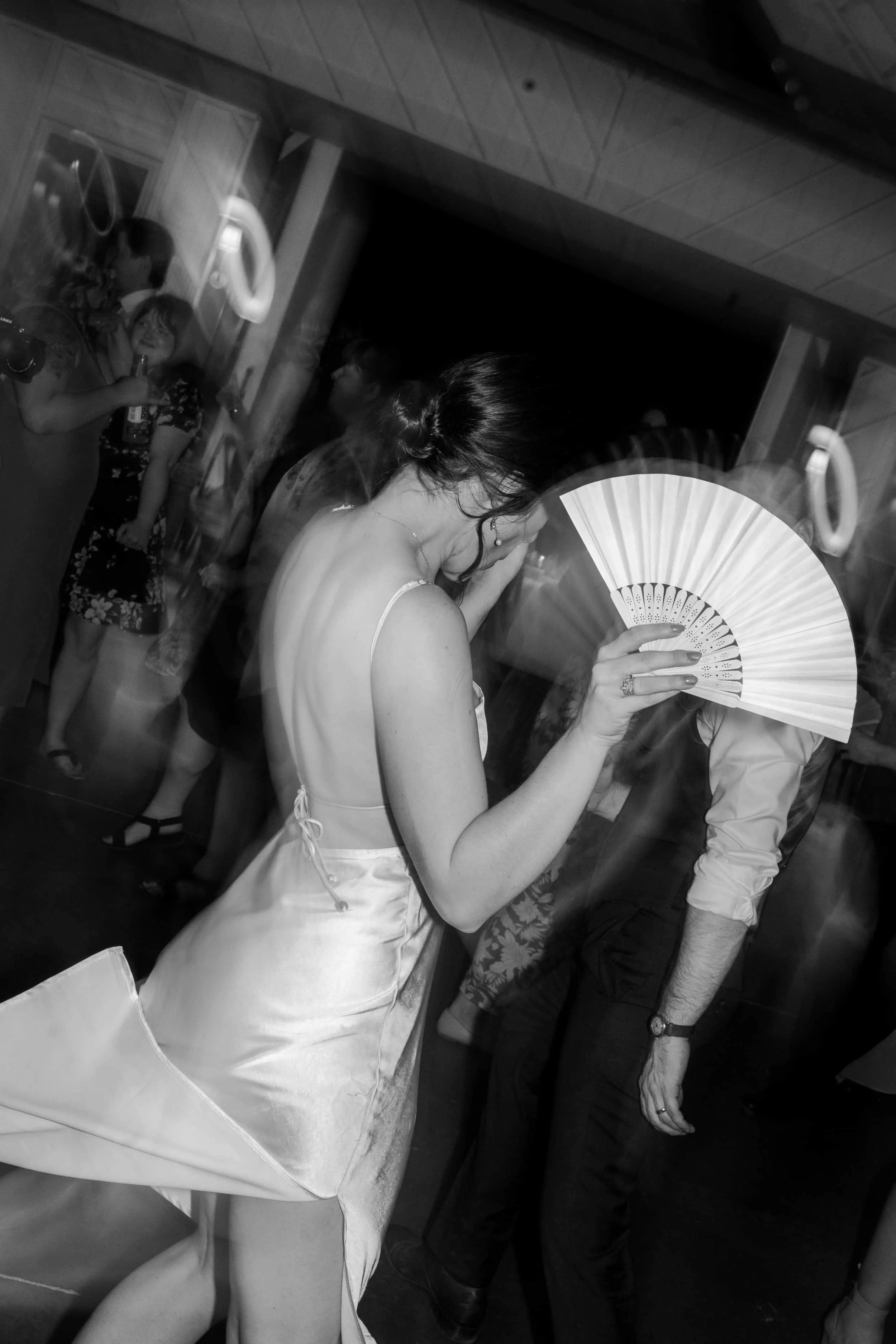 A woman in a satin dress holding a fan at a party or dance event, with other people in the background.