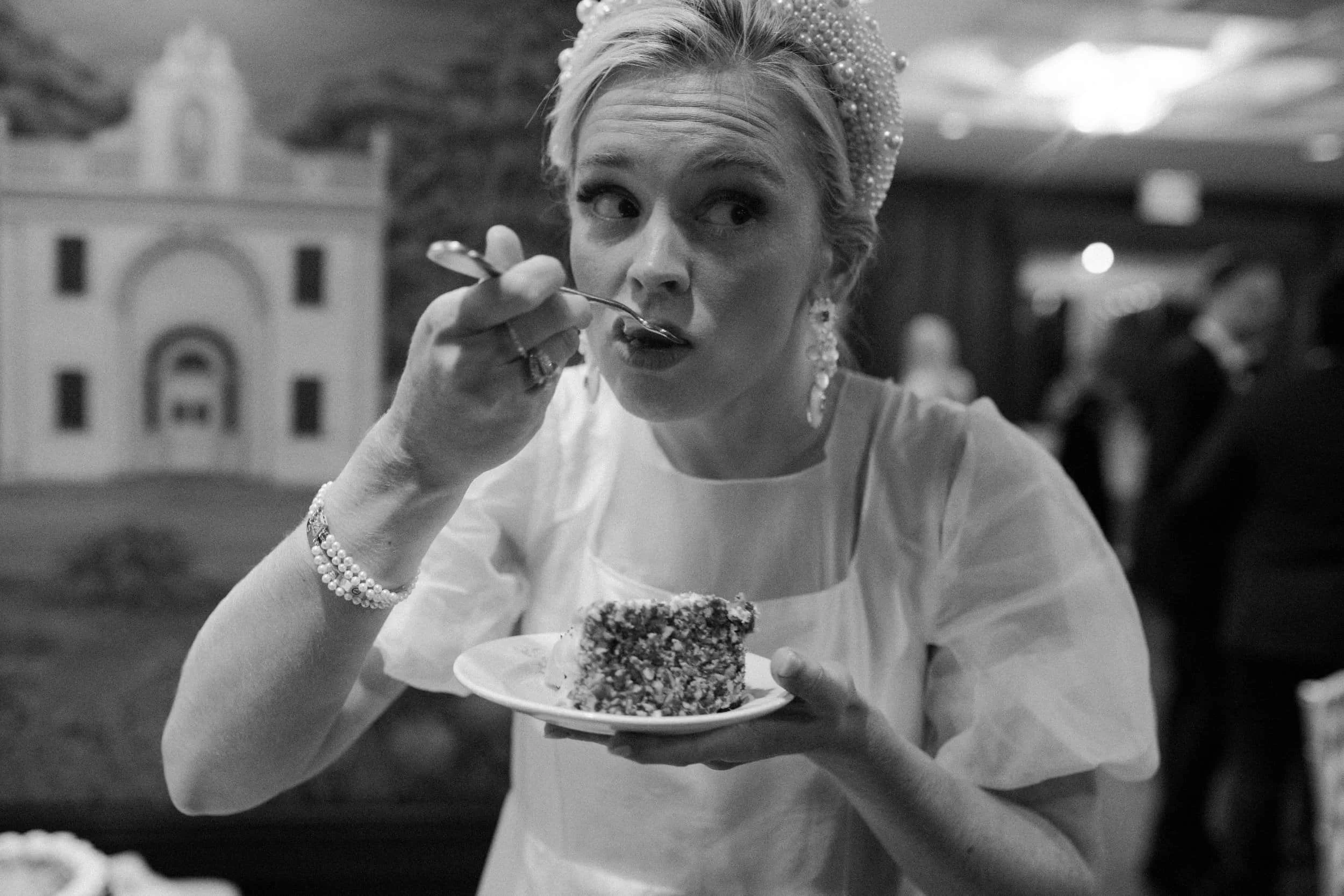 A woman wearing a pearl headband and earrings is eating a slice of cake with a fork while holding a small plate. She is dressed in a light-colored dress and is wearing a beaded bracelet. The background shows a blurred interior setting with other peop