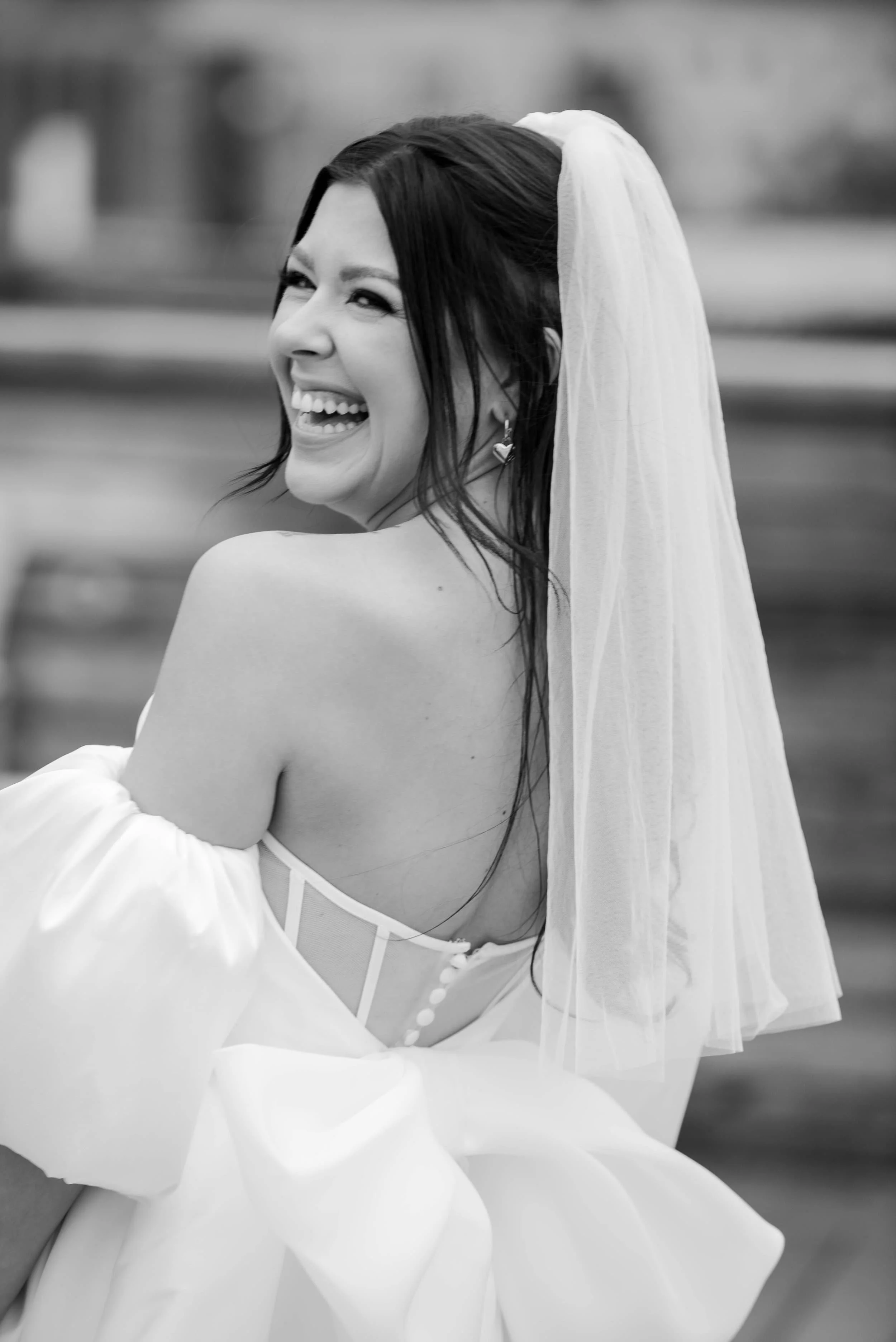 A woman in a wedding dress and veil, smiling and looking over her shoulder outdoors.