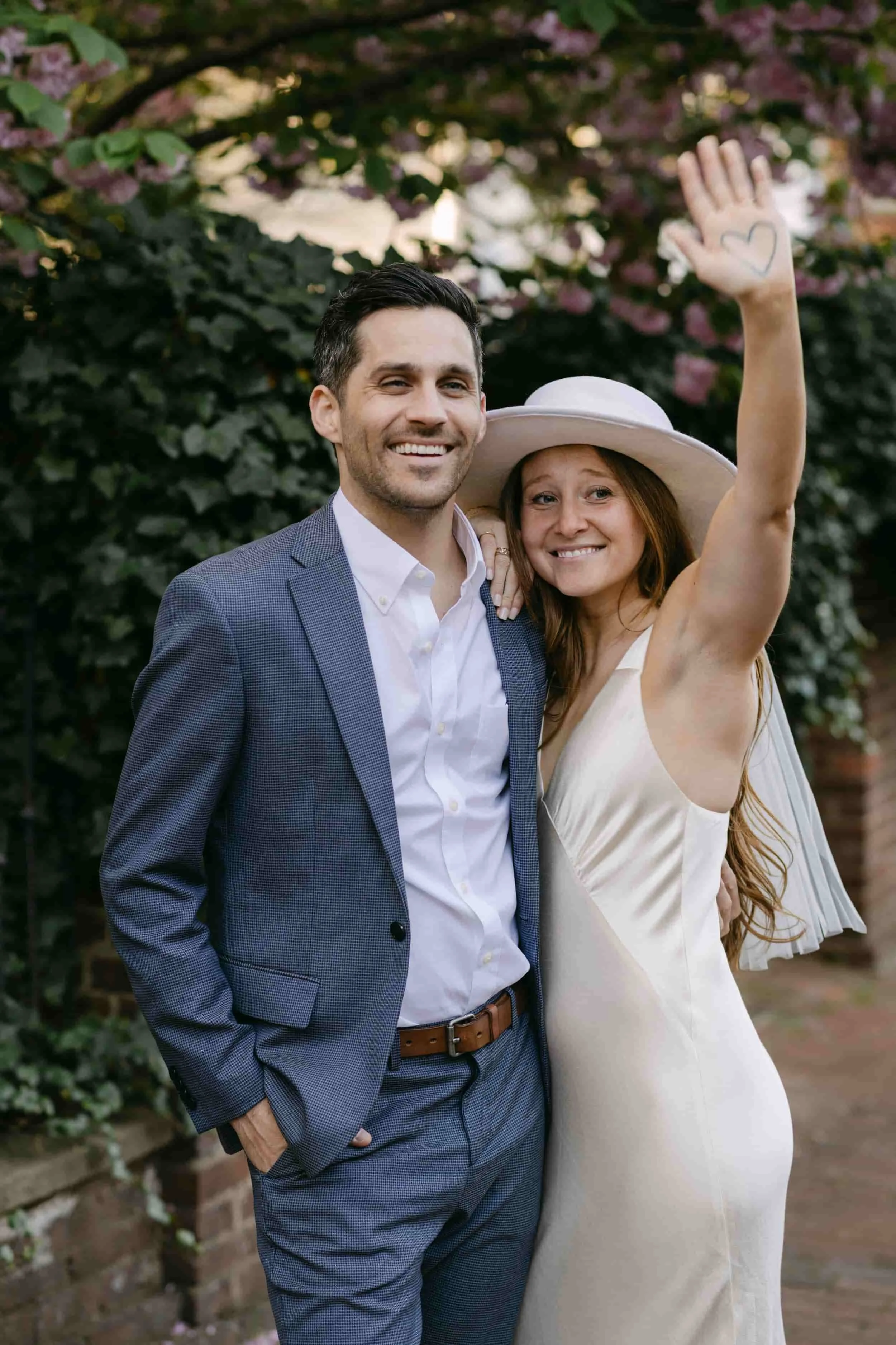 A smiling couple standing outdoors with blooming pink flowers and greenery in the background. The woman is wearing a white dress and a large white hat, raising her hand with a heart drawn on it. The man is dressed in a gray suit jacket, white shirt, 