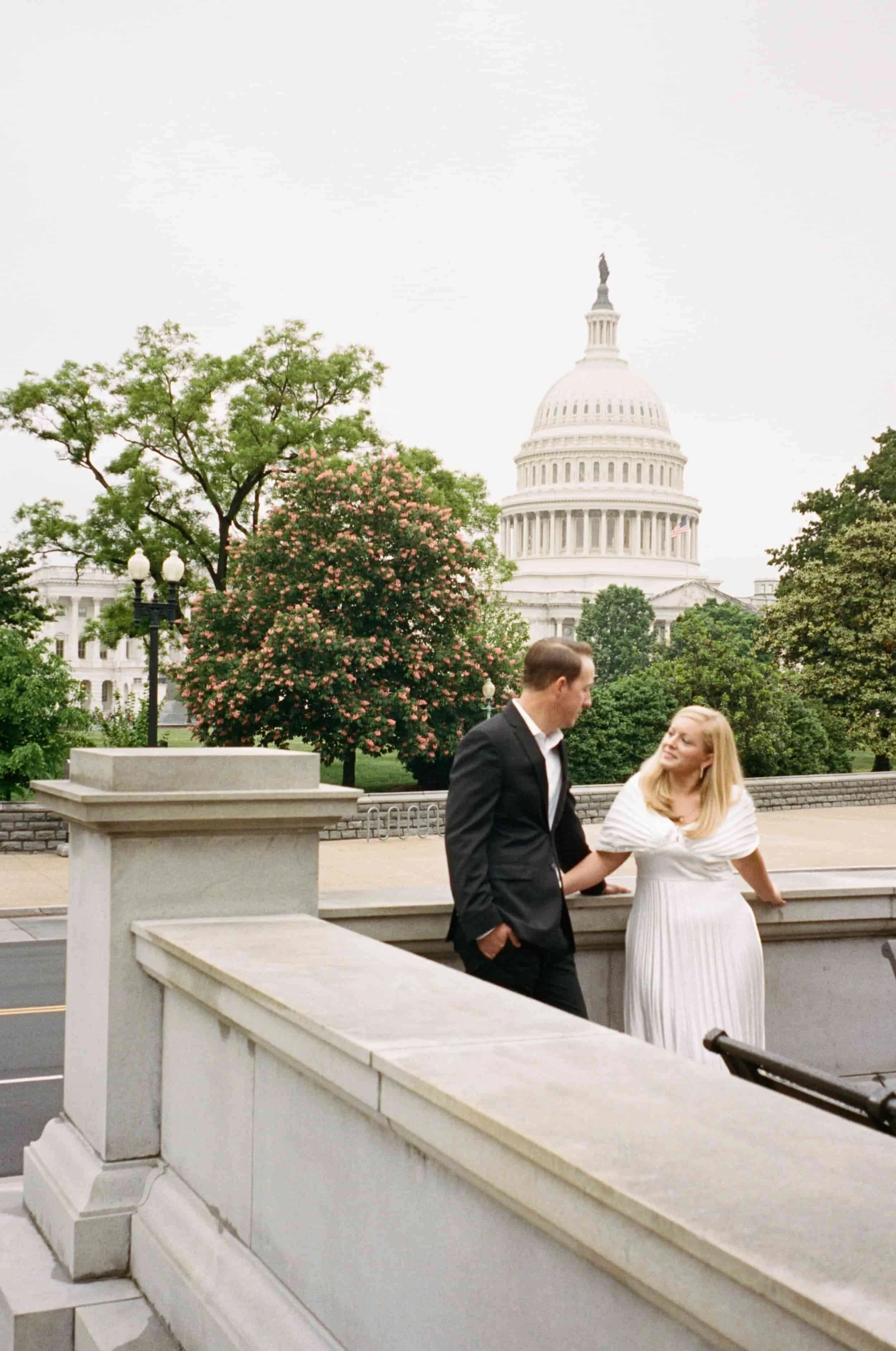 A couple in formal attire standing on a white stone balcony outside the U.S. Capitol building, with trees and a pink flowering bush in the background.