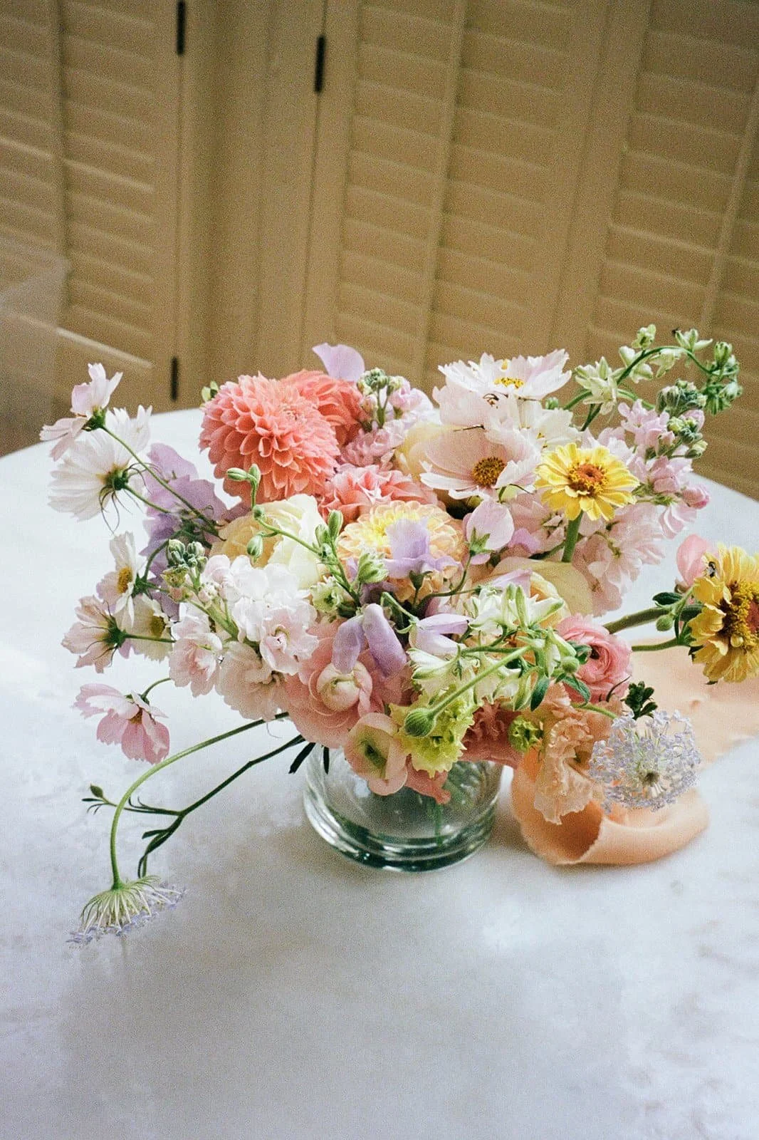 A glass vase filled with a colorful arrangement of pink, white, yellow, and purple flowers on a white table with beige window shutters in the background.