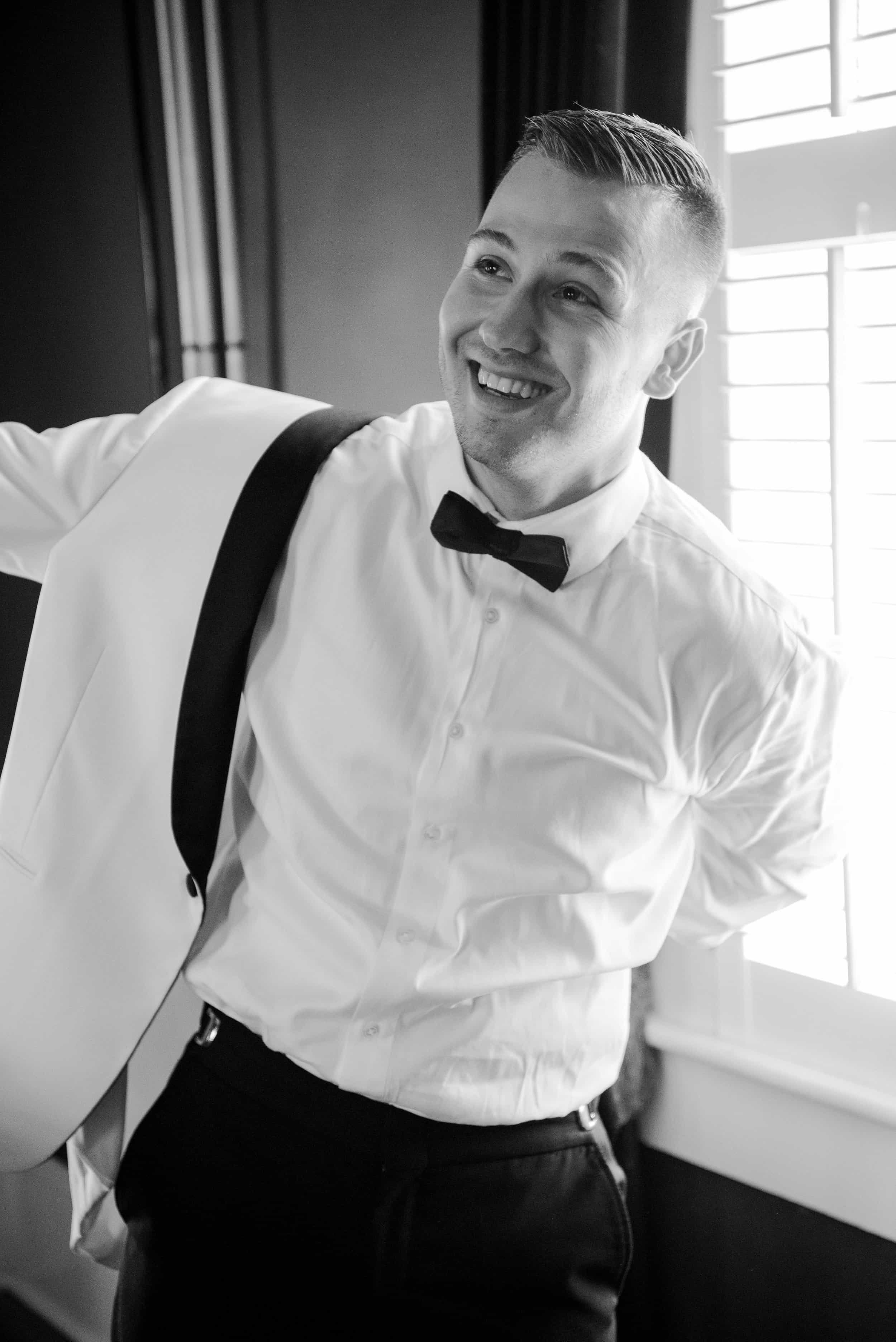 A young man dressed in formal attire with a bow tie, white shirt, and suspenders, smiling and posing indoors near a window with blinds.