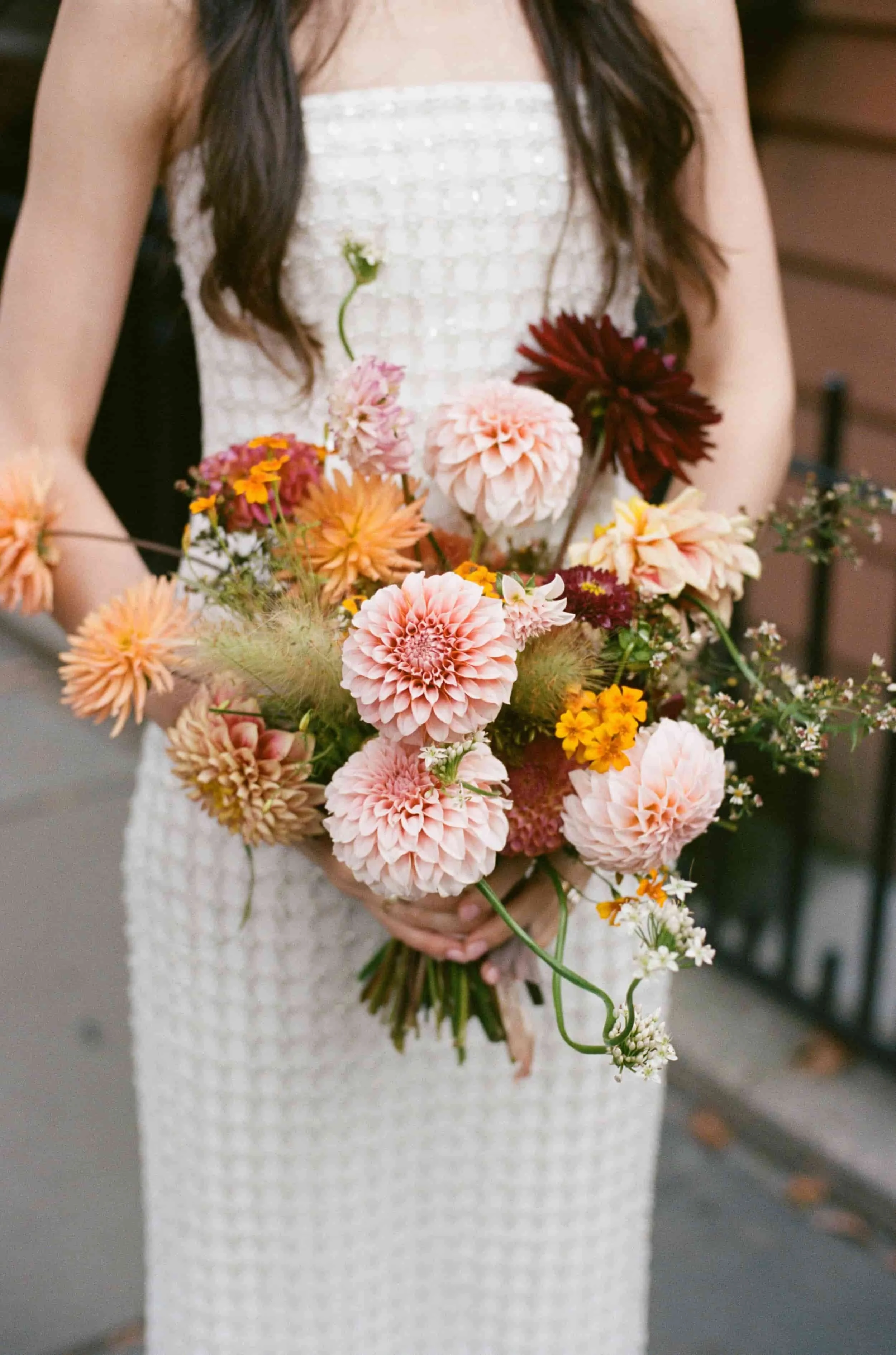 A person wearing a white textured dress holding a large bouquet of pink, peach, orange, and burgundy flowers including dahlias and other mixed blooms.
