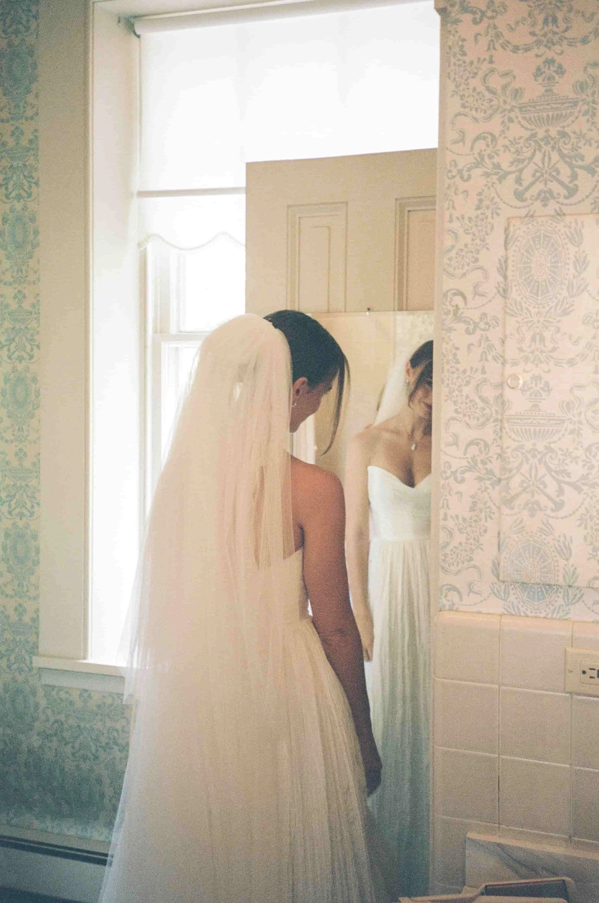 A bride in a wedding dress with a veil looking into a mirror in a light-filled room.