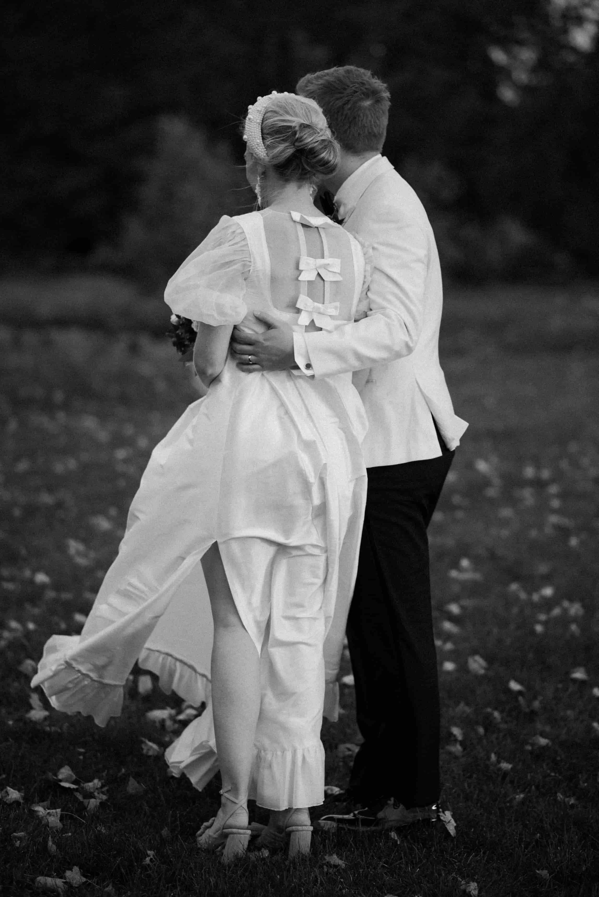 A couple dressed in wedding attire, embracing outdoors on a grassy area, in black and white.