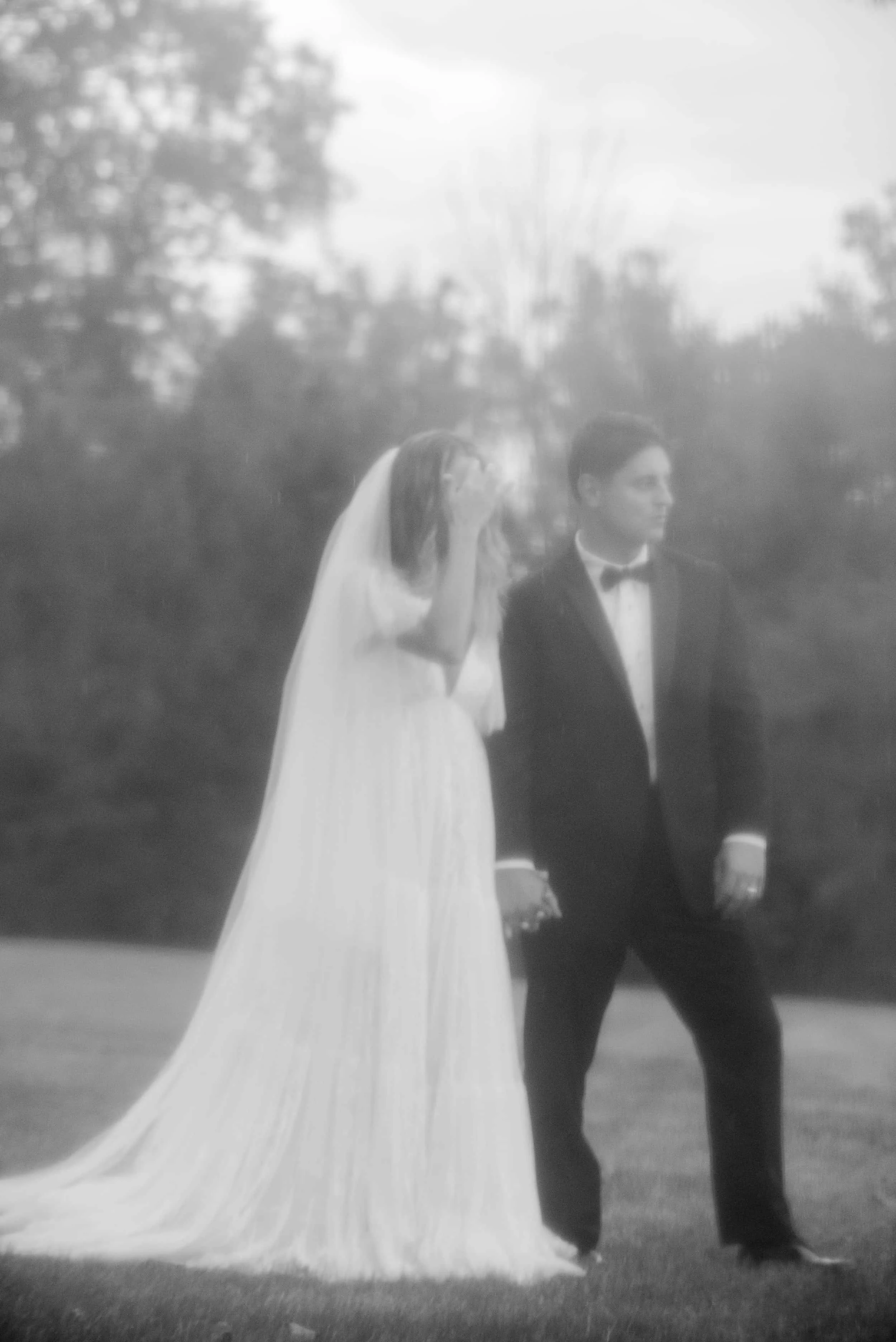 Black and white photo of a bride and groom outdoors during a wedding ceremony. The bride is wearing a long gown with a veil, and the groom is dressed in a tuxedo. They are walking hand in hand on a grassy area with trees in the background.