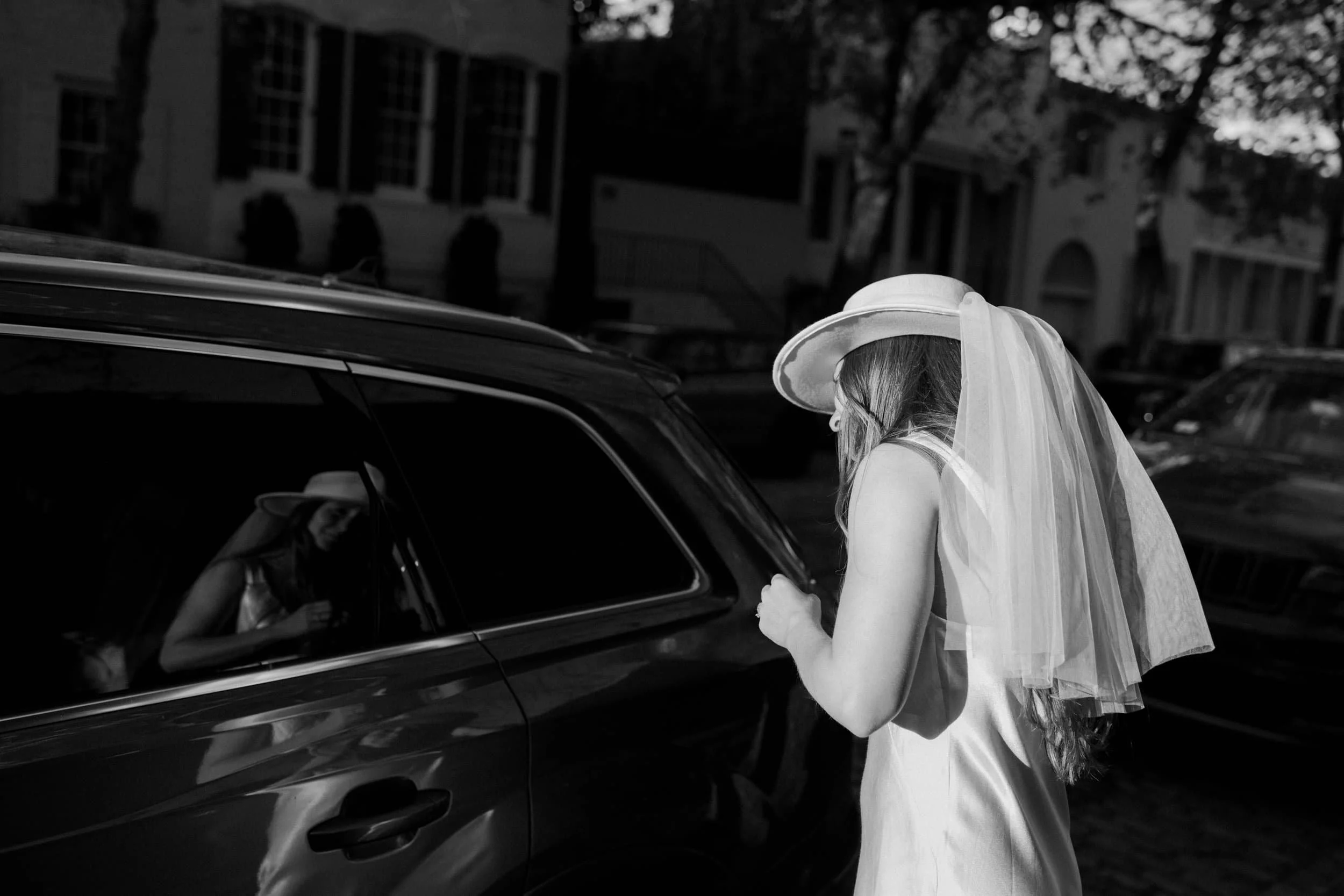 A woman in a wedding dress and hat with a veil is standing next to a car, looking at her reflection in the window.