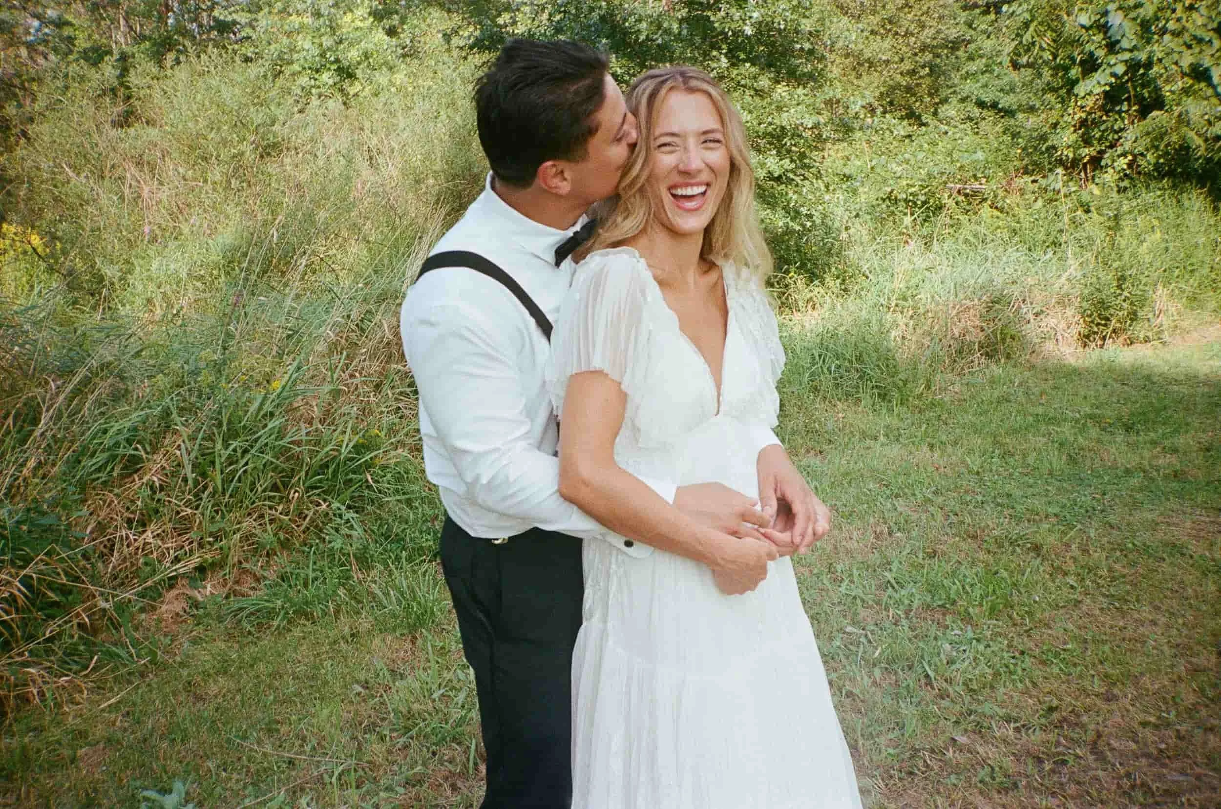 A joyful couple dressed in wedding attire, standing outdoors on a grassy area with bushes and trees in the background. The man is kissing the woman on the cheek, and she is smiling.