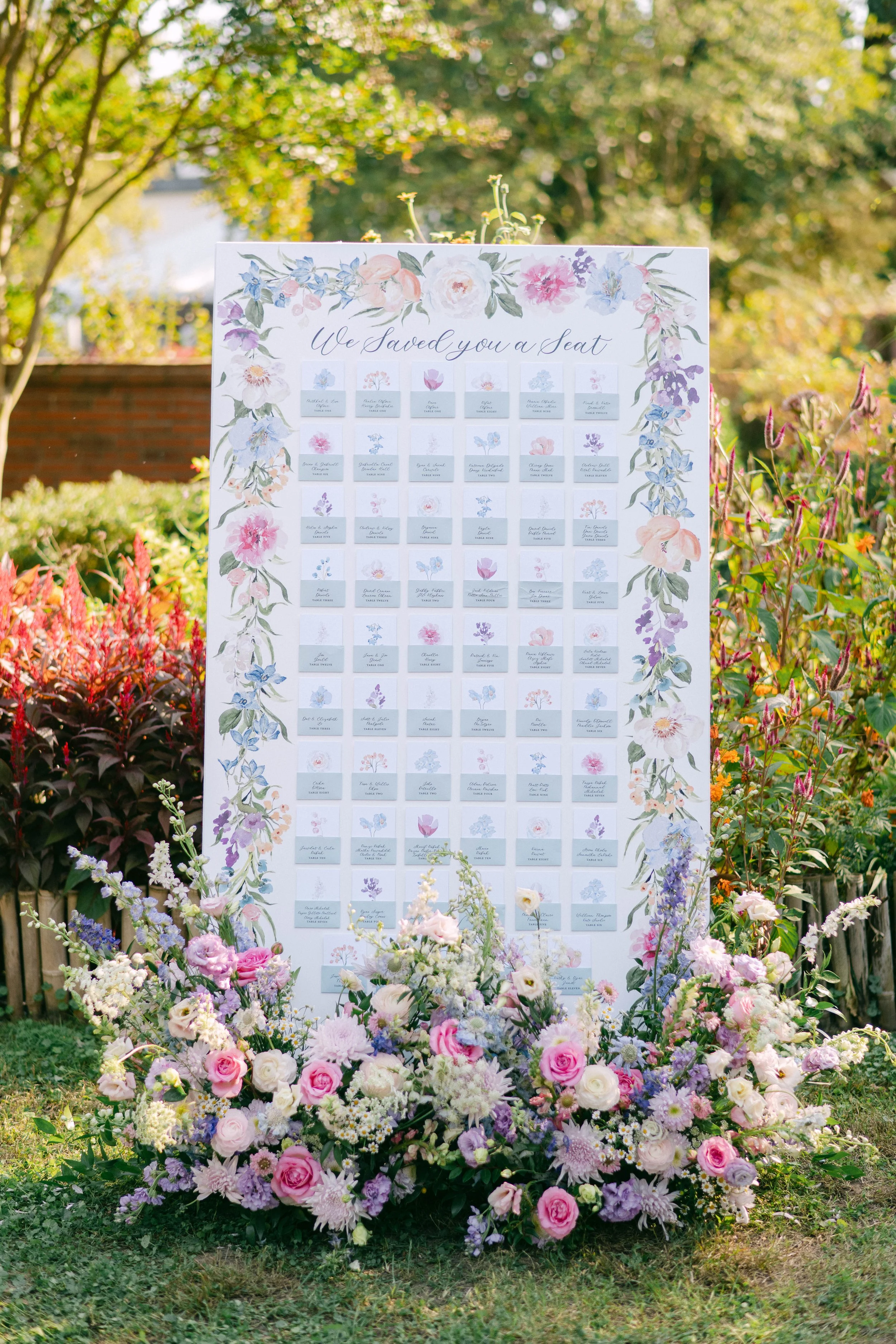 A floral wedding seating chart with small individual cards attached, surrounded by an arrangement of pink and purple flowers, placed outdoors with greenery and trees in the background.