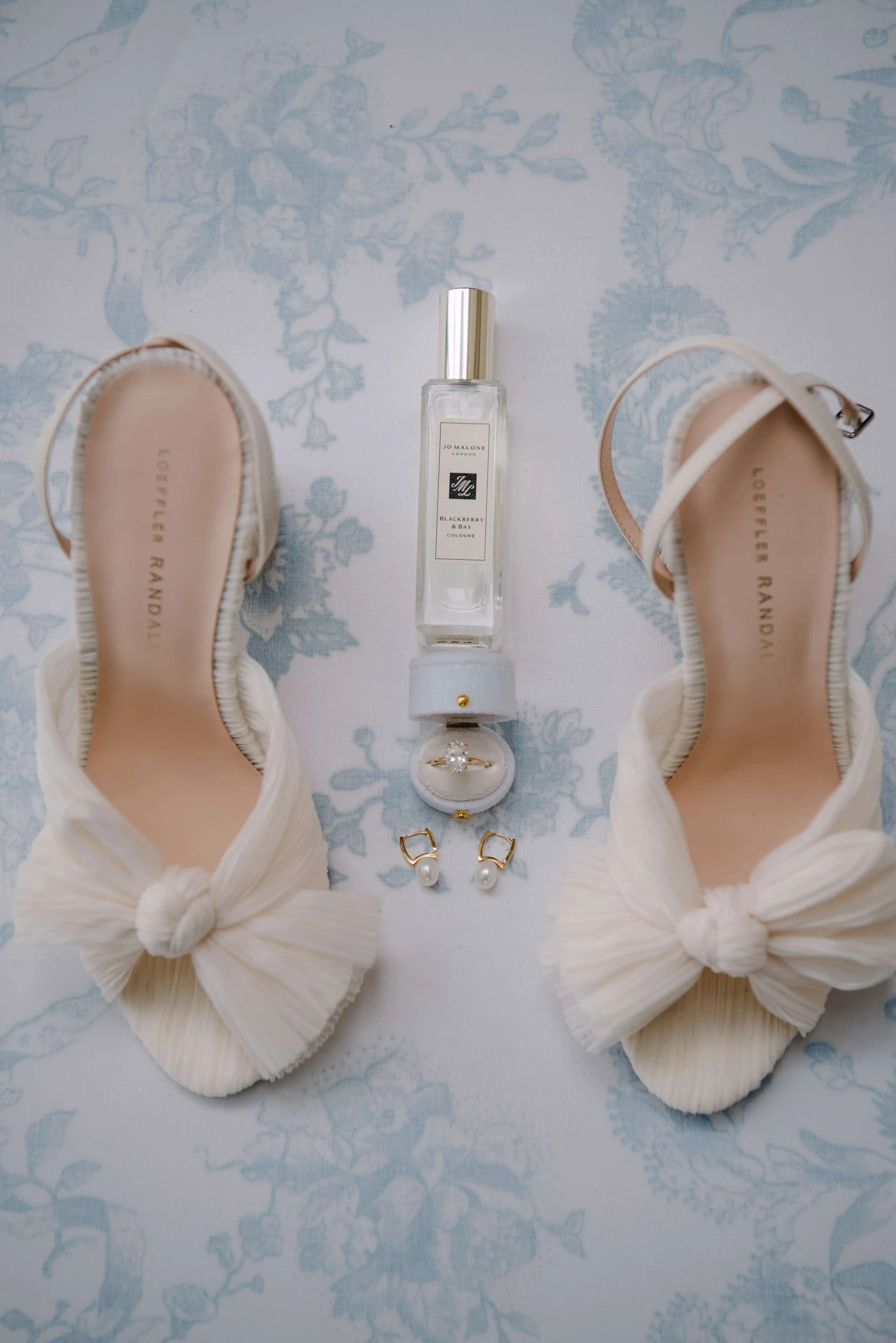 Pair of beige shoes with large bows, a perfume bottle, a jewelry box with a ring, and a pearl earring with a gold clasp displayed on a blue floral patterned fabric.