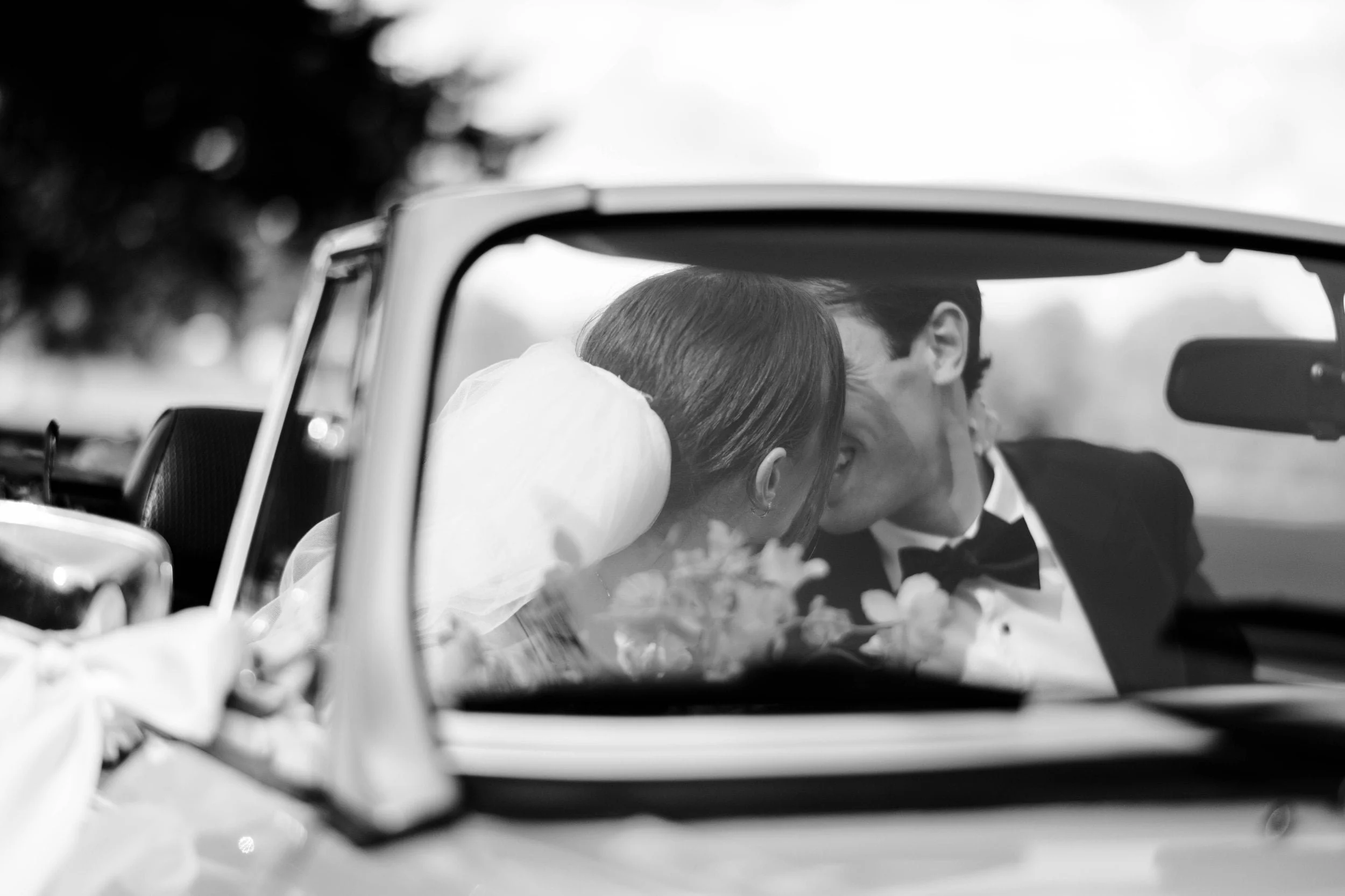 A black-and-white photo of a couple dressed in wedding attire leaning in for a kiss inside a vintage convertible car.