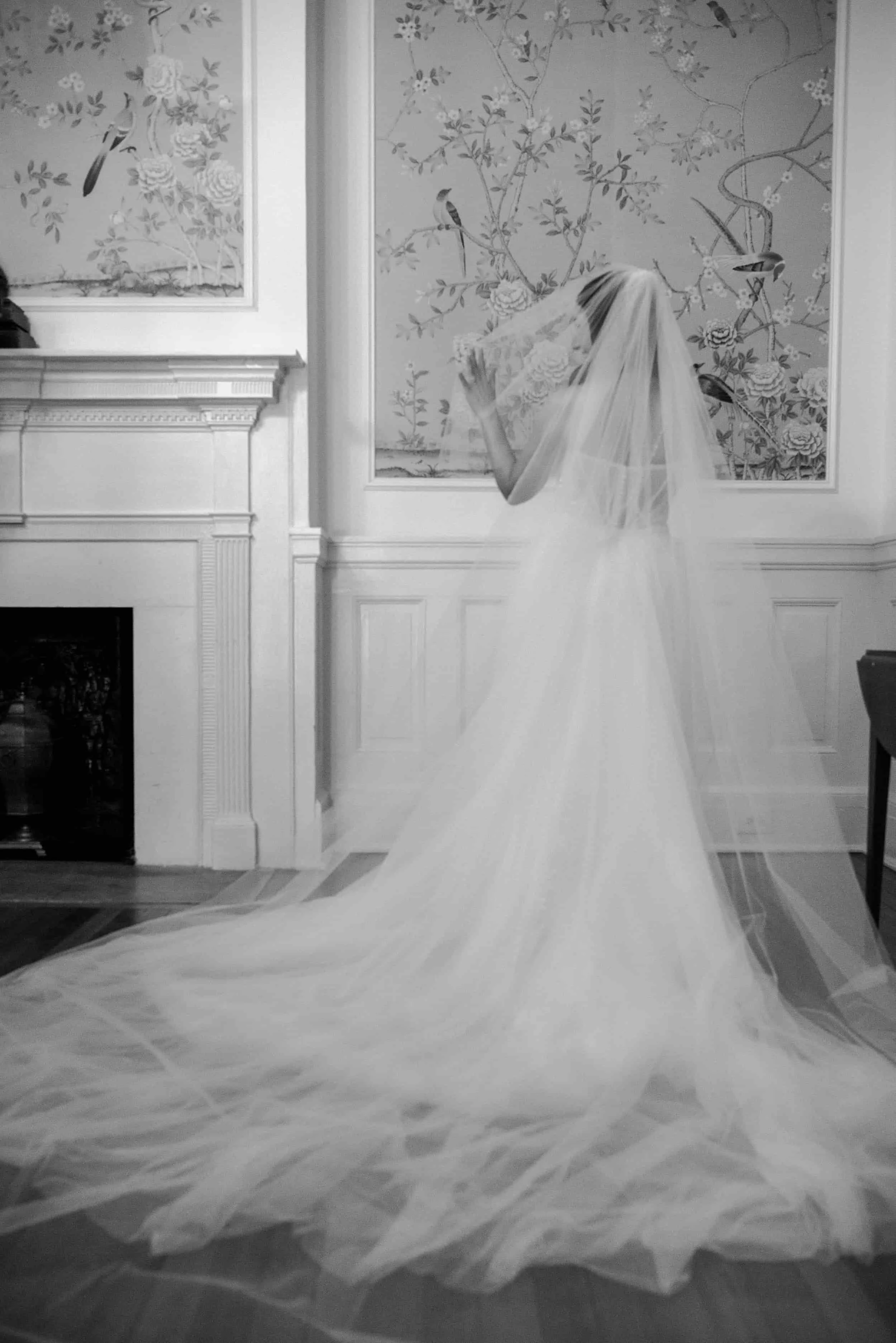 A woman in a wedding dress with a long veil, standing indoors near a decorated wall and fireplace, holding her veil near her face.