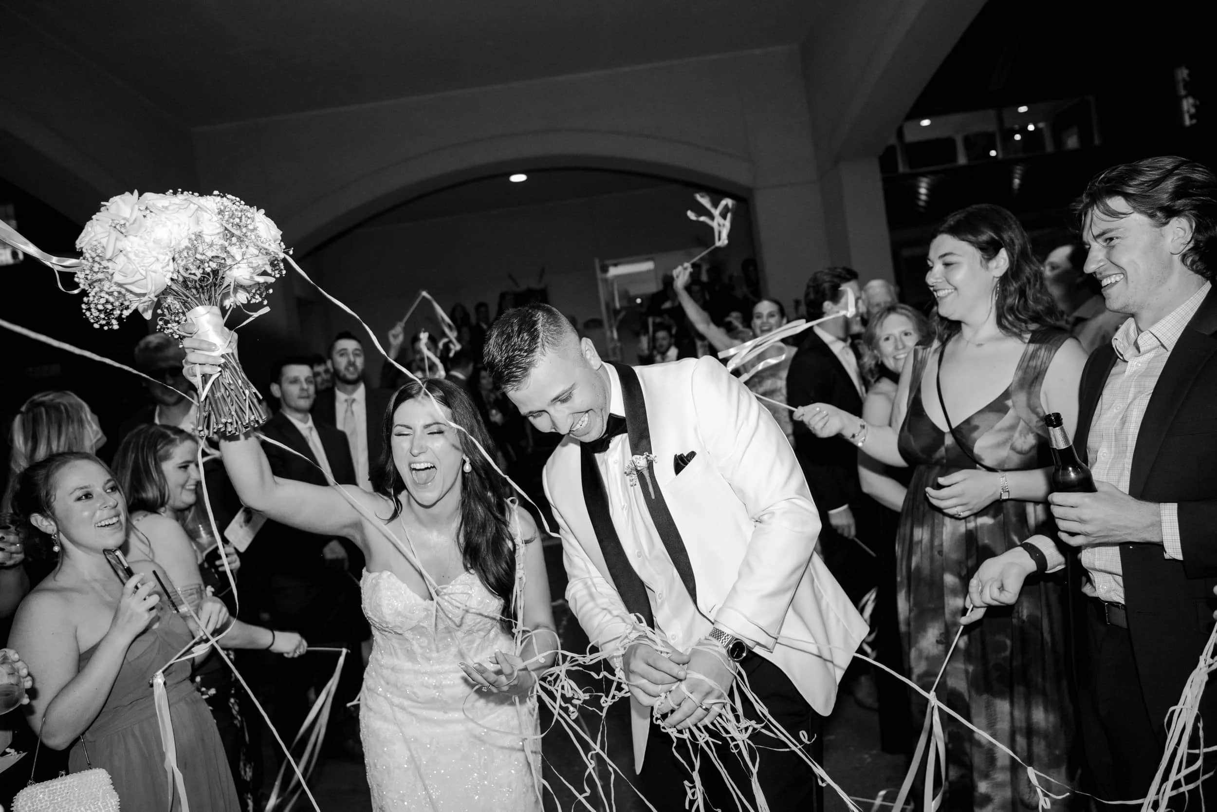 A wedding celebration with a bride and groom at the center, surrounded by friends and family. The bride is holding a bouquet and smiling, while the groom is laughing and holding a ribbon. People around are joyfully participating in a party, with some