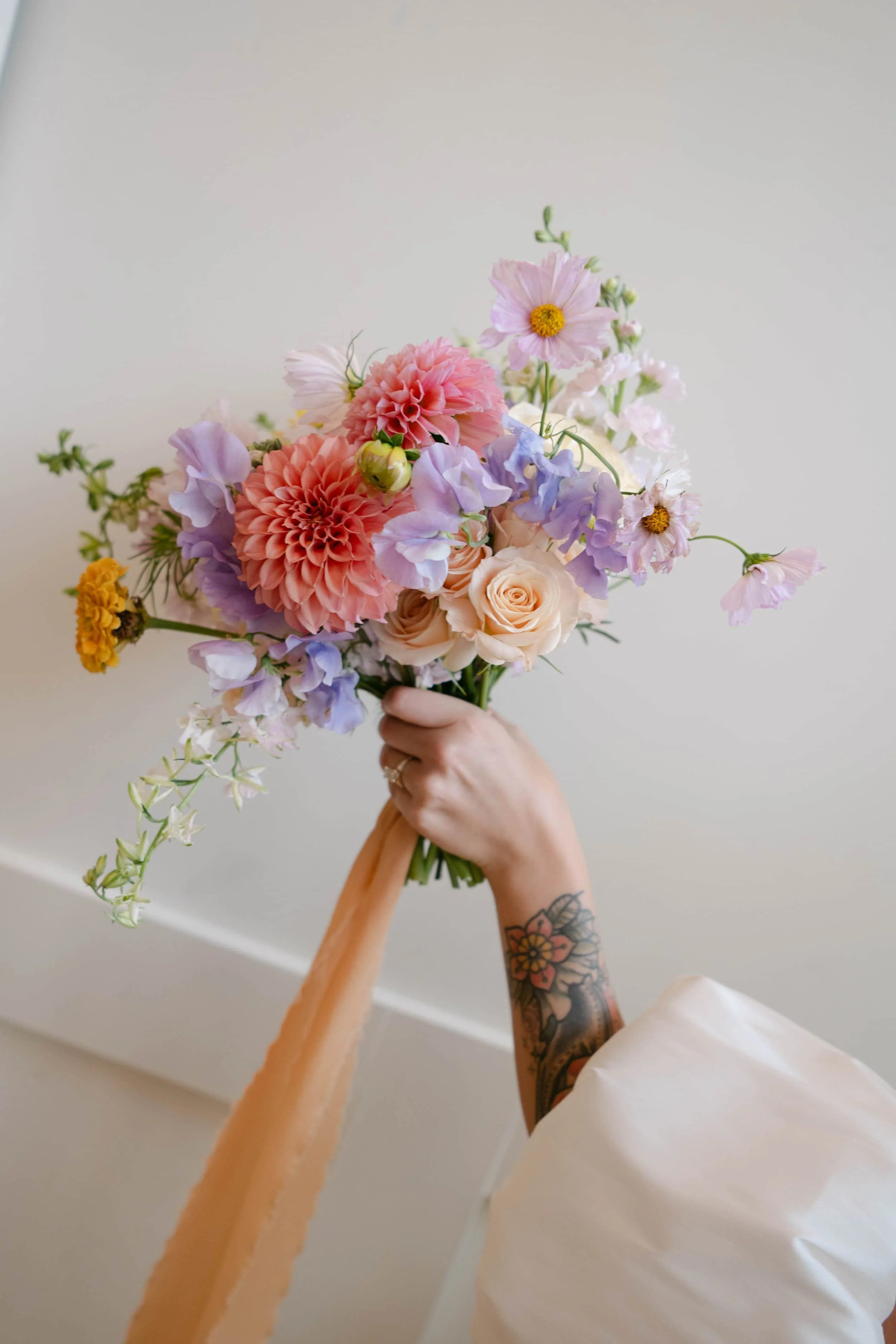 Person holding a bouquet of mixed flowers including roses, dahlias, and other blooms against a plain white background.