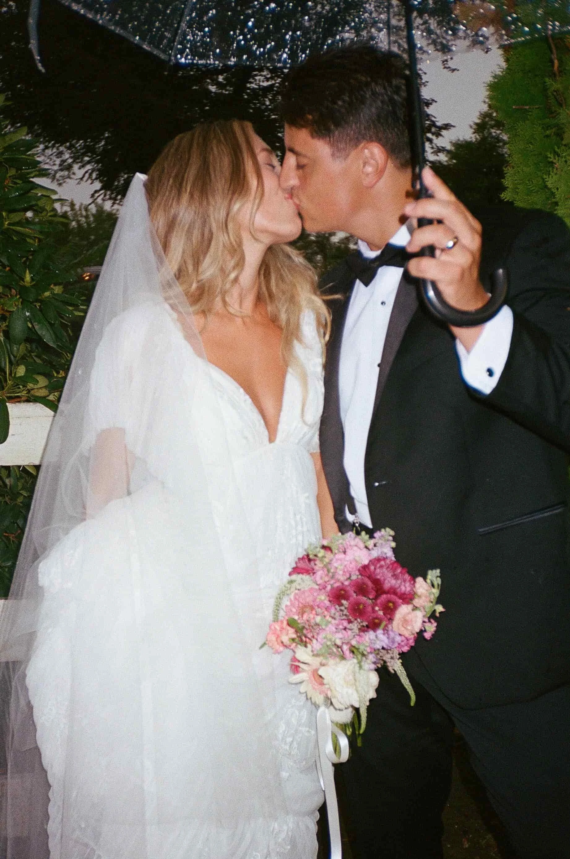 A bride in a wedding dress and a groom in a tuxedo kiss under an umbrella, with the bride holding a pink and purple bouquet of flowers.