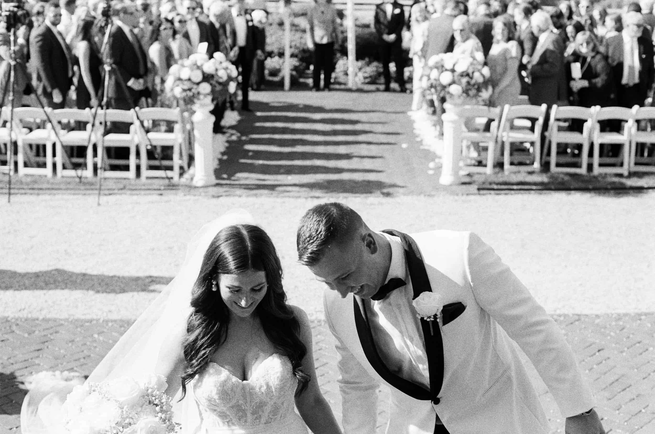Black and white photo of a bride and groom at their outdoor wedding, leaning down side by side, smiling. The bride wears a strapless wedding gown with a veil and holds a bouquet. The groom is in a tuxedo with a bow tie and boutonniere. In the backgro