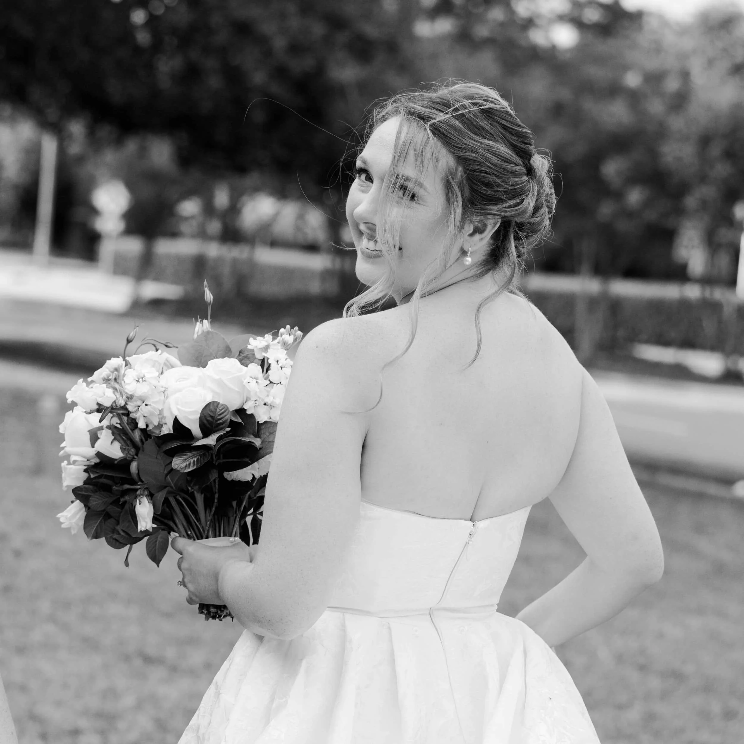 A smiling woman in a strapless dress holding a bouquet of flowers outdoors.