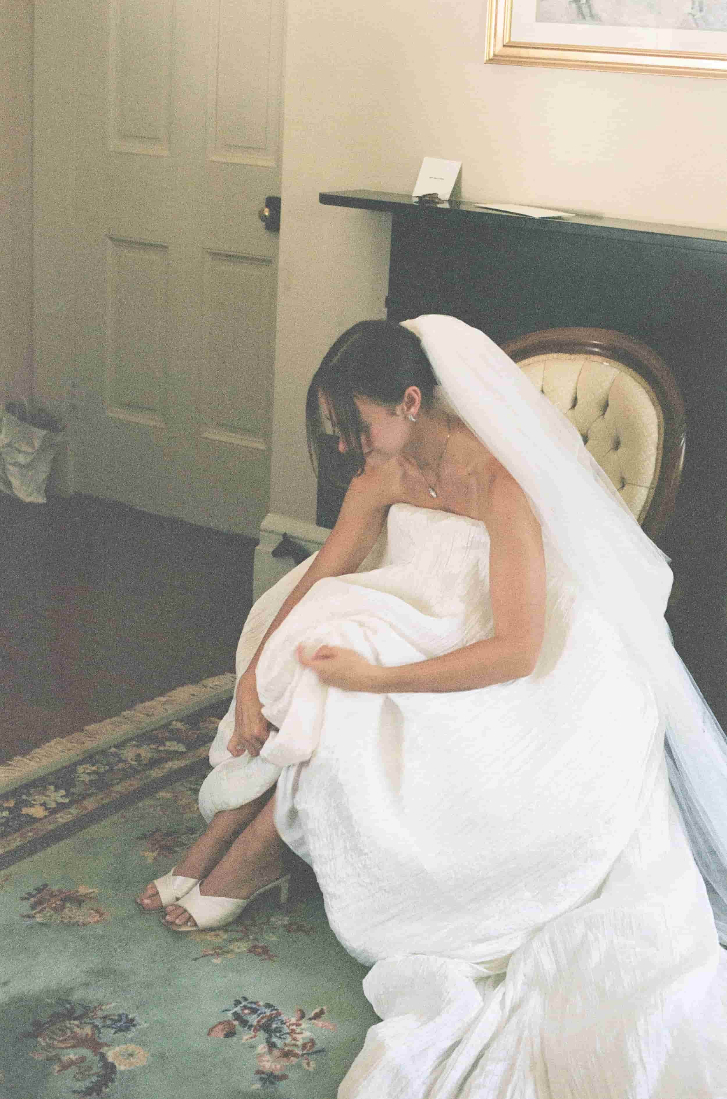 A bride dressed in a white wedding gown and veil sitting on a bed, putting on her stocky.