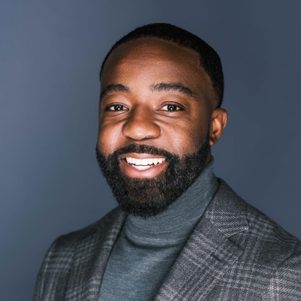 Headshot of a man smiling, wearing a business suit and a turtleneck, against a solid dark background.