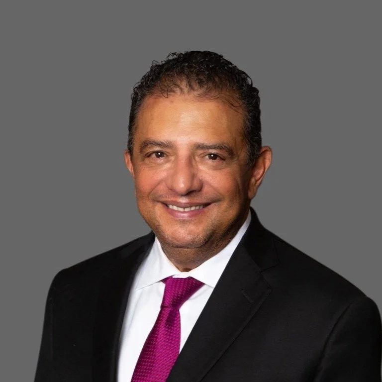 Portrait of a middle-aged man in a black suit with a white shirt and purple tie, smiling against a gray background.