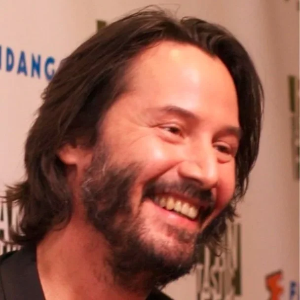 Headshot of Keanu Reeves smiling away from the camera.