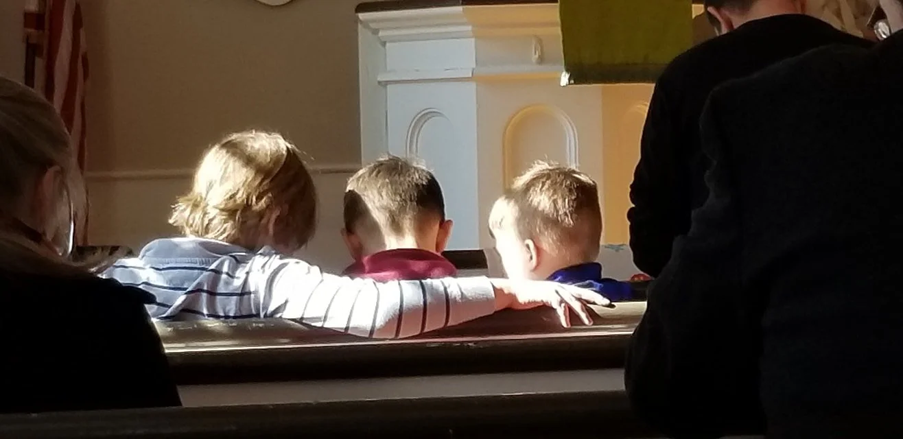 Three children sitting closely on a church pew, with one putting an arm around the others.