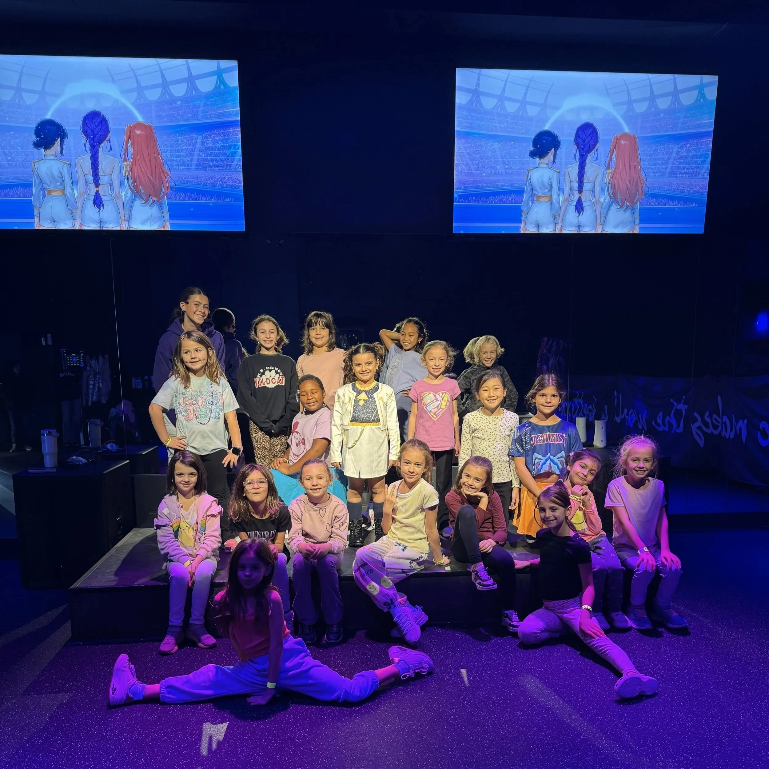 Group of young girls and a woman in a dark indoor setting with two screens above them displaying animated characters watching a sports event.