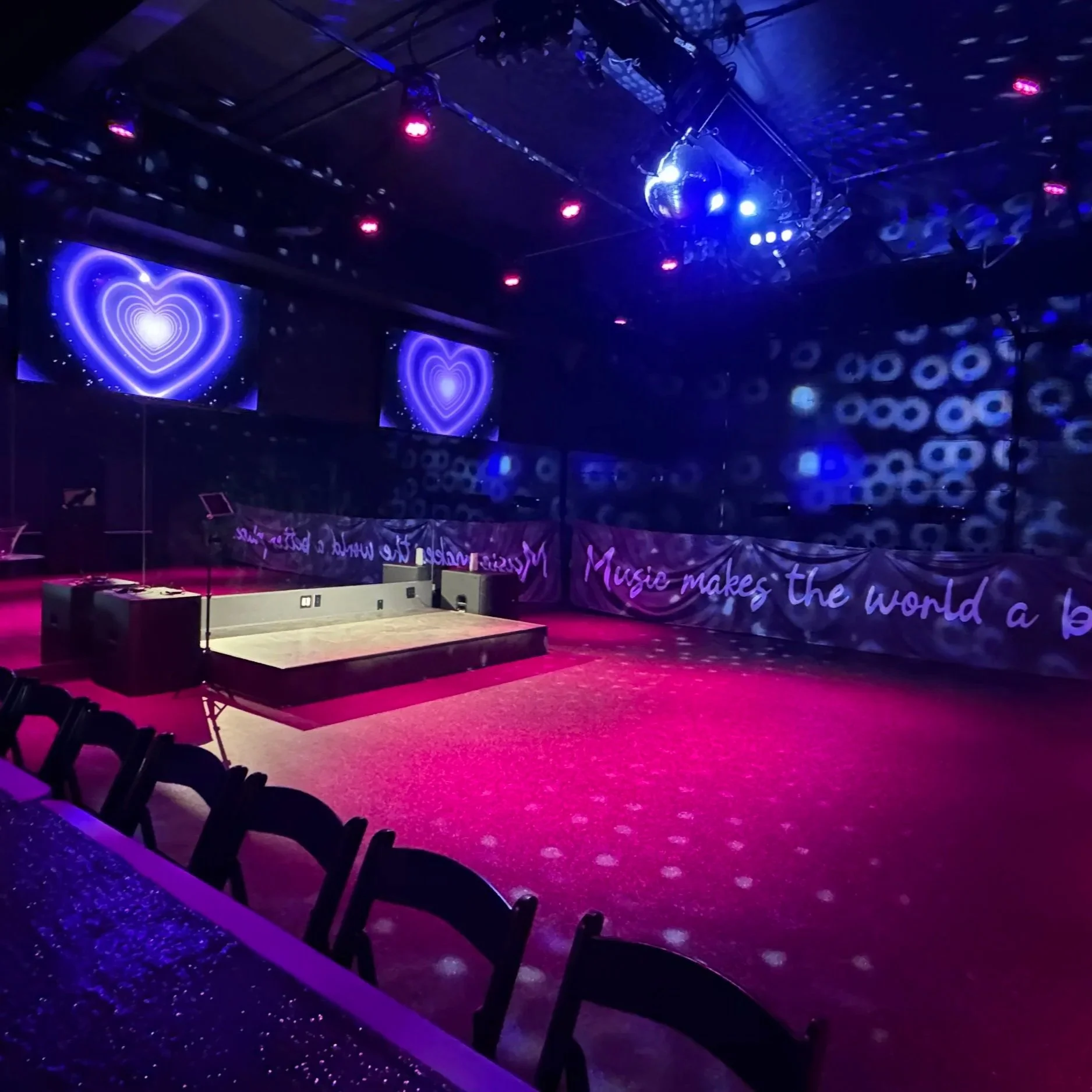 Empty stage in a dimly lit nightclub or event venue with purple and pink lighting, two large screens displaying glowing hearts, a banner with the phrase 'Music makes the world a better place,' and a few chairs along a decorated wall.
