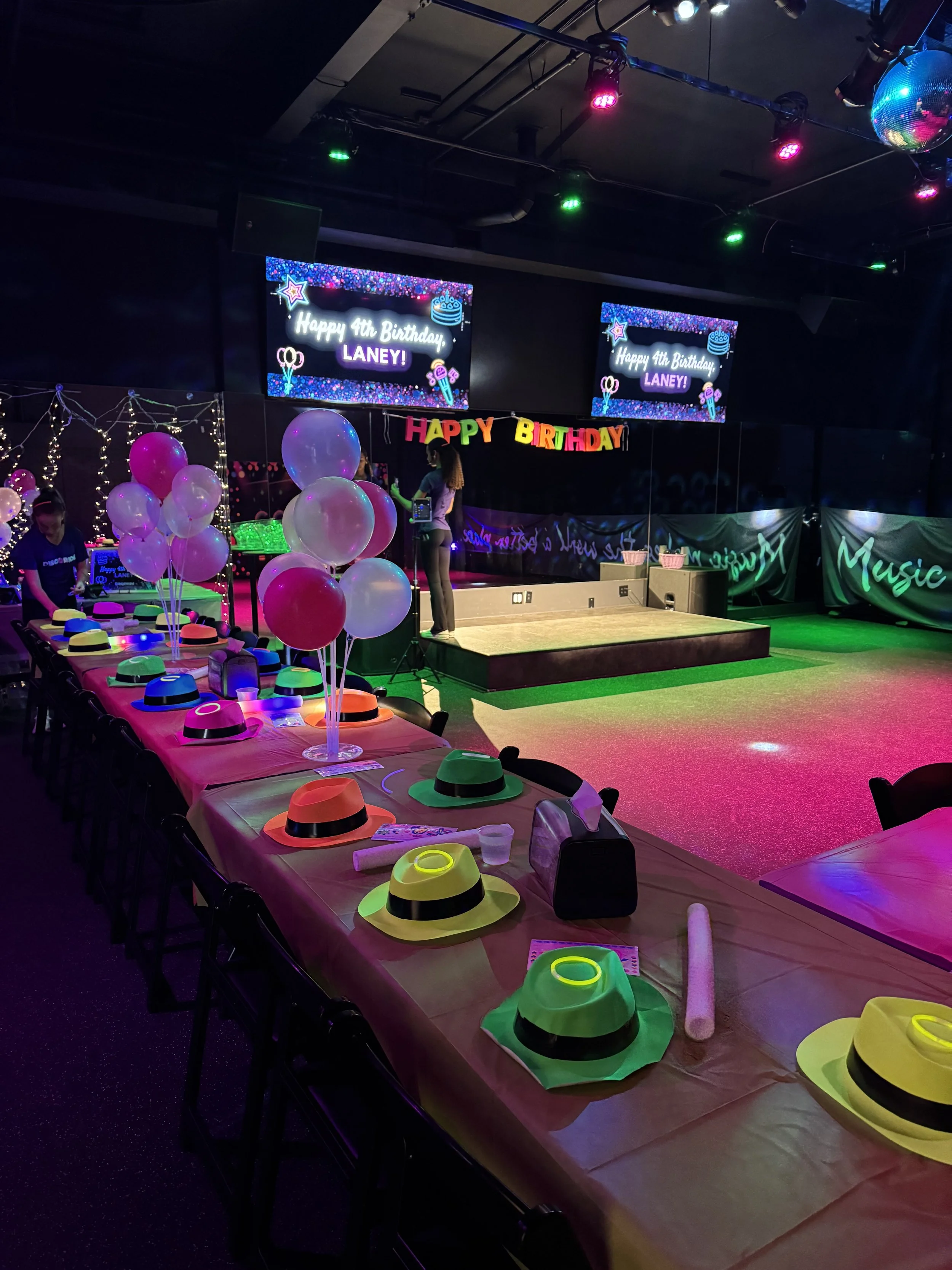 Colorful birthday party setup with balloons, hats, and decorations. A large banner says 'Happy 4th Birthday, Laney!' and birthday hats are on a long table. The stage is decorated for celebration with digital screens and festive lighting.