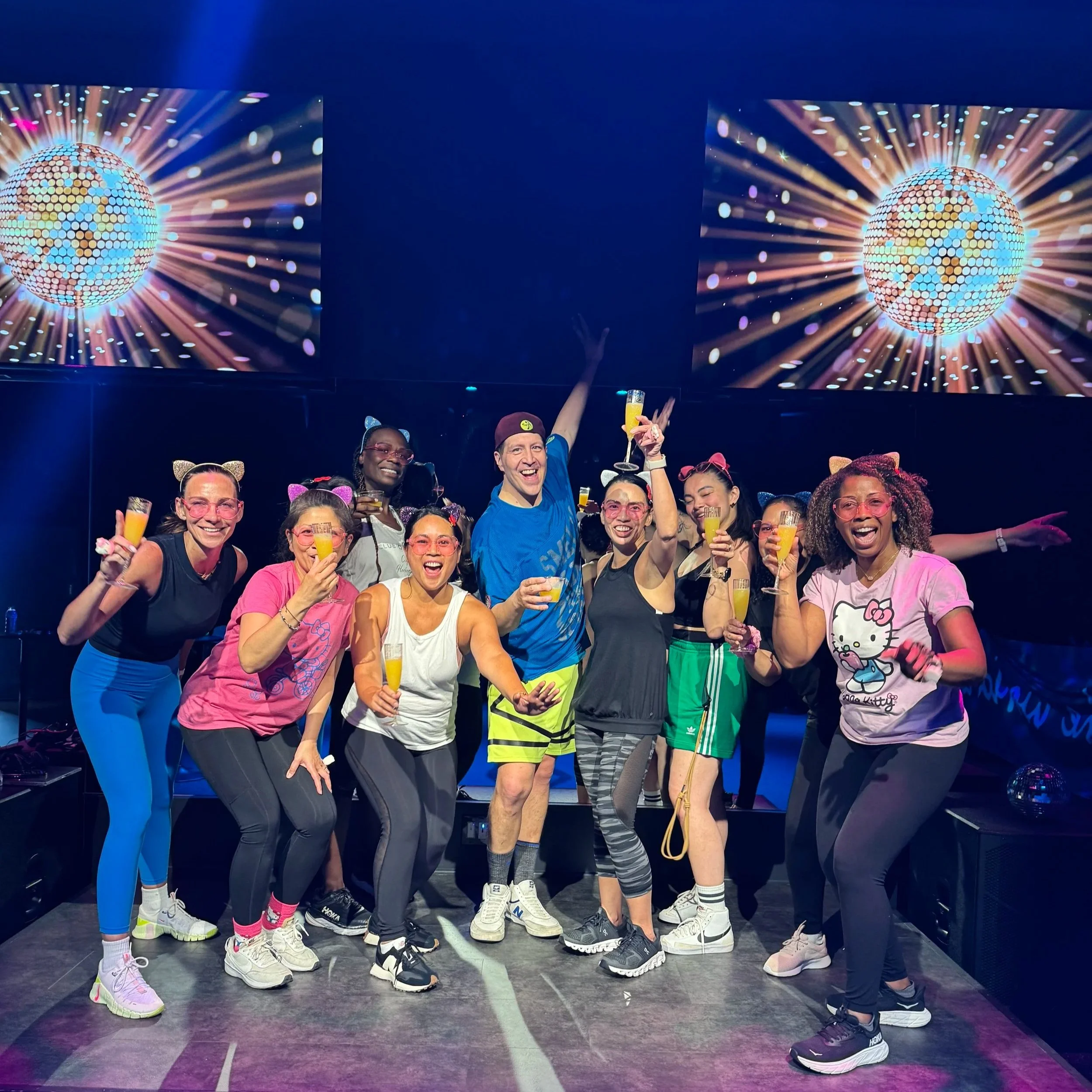 Group of people on a stage celebrating with drinks at a party, with colorful disco-themed background screens showing a globe with light rays.