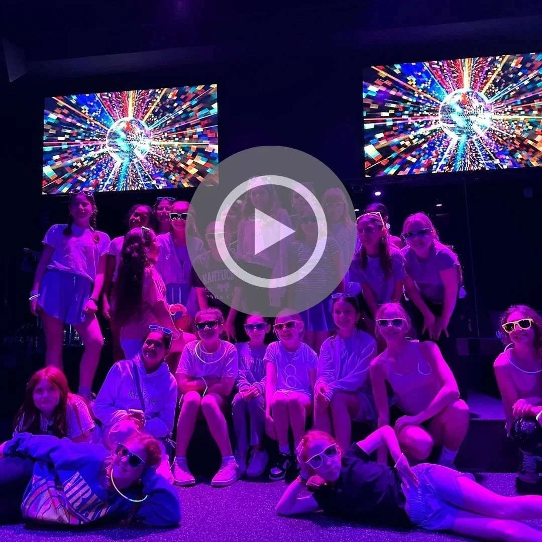 Group of children wearing sunglasses in a dark room with colorful LED screens showing a digital globe and bursting light patterns.