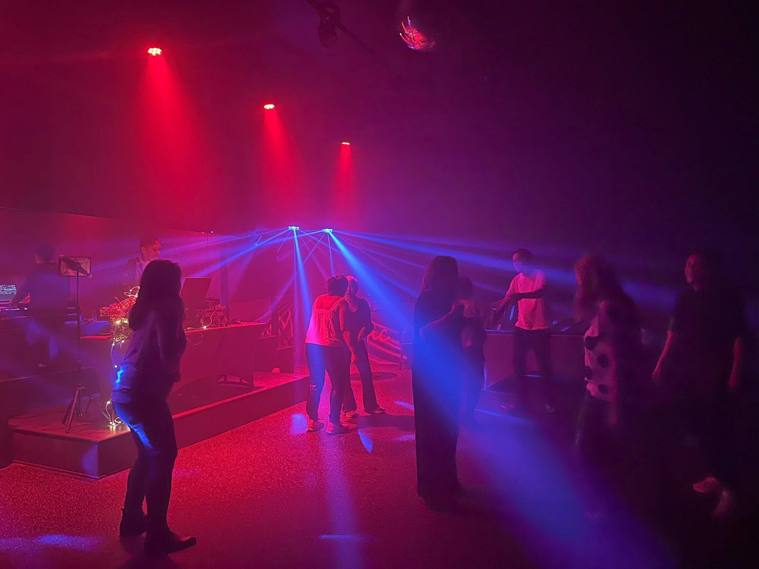 People dancing and socializing at a nightclub with colorful lights and a DJ setup.