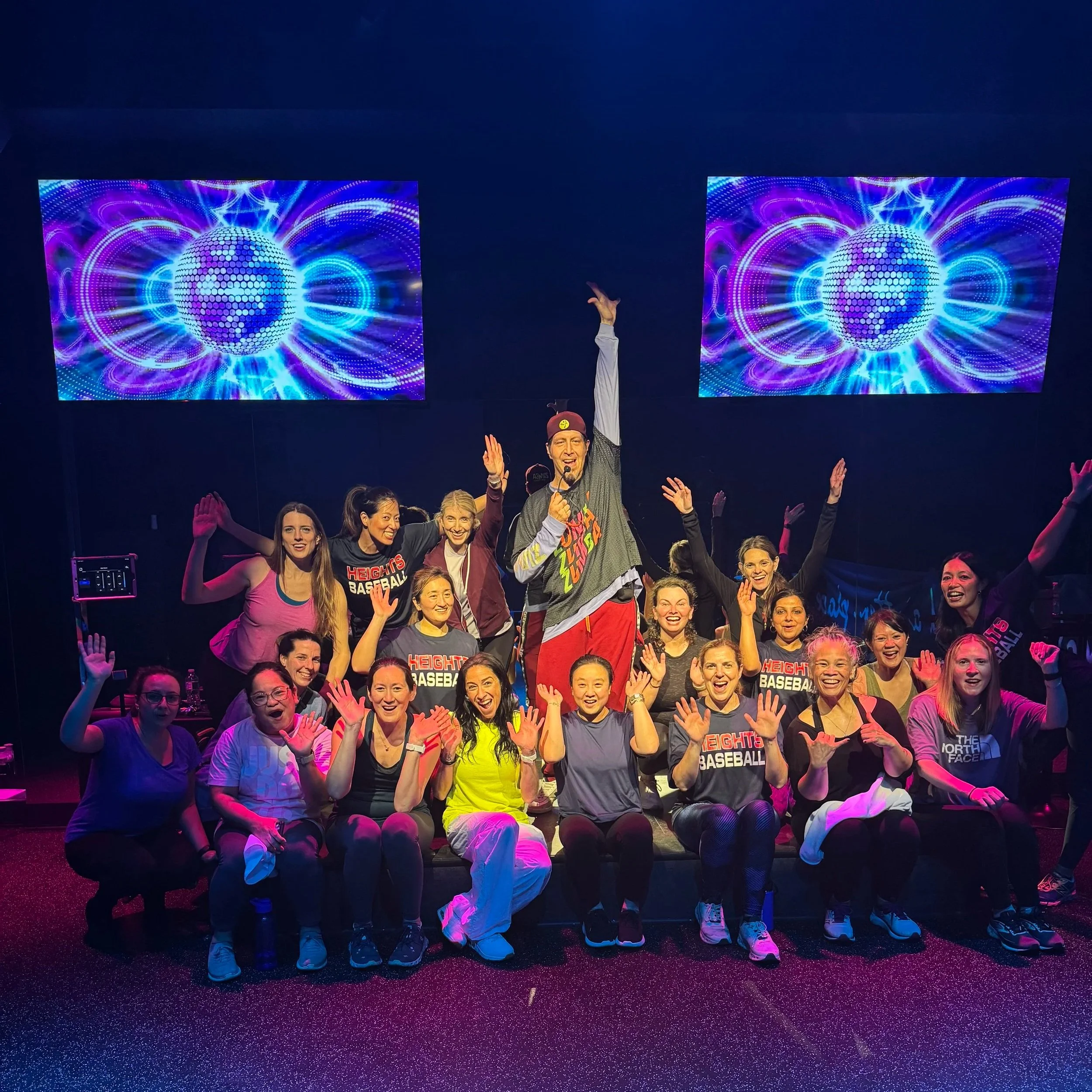 Group of people on stage celebrating with a man holding a microphone, surrounded by colorful lights and two large screens displaying digital graphics in a dark room.