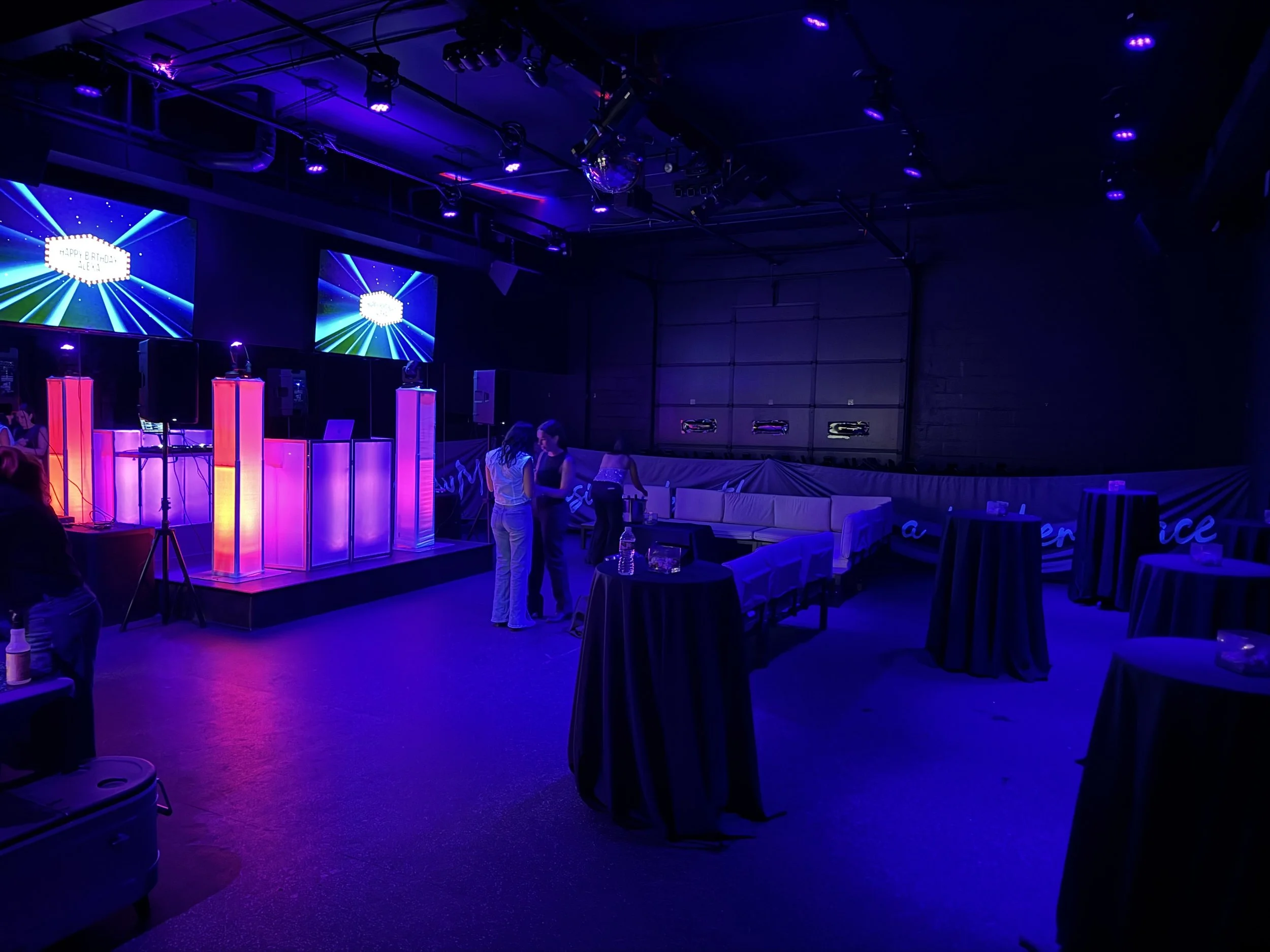 Empty event space with dim blue and purple lighting, a stage with large vertical color-changing light bars, two screens showing a celebratory message, and a few people standing and talking near the stage.