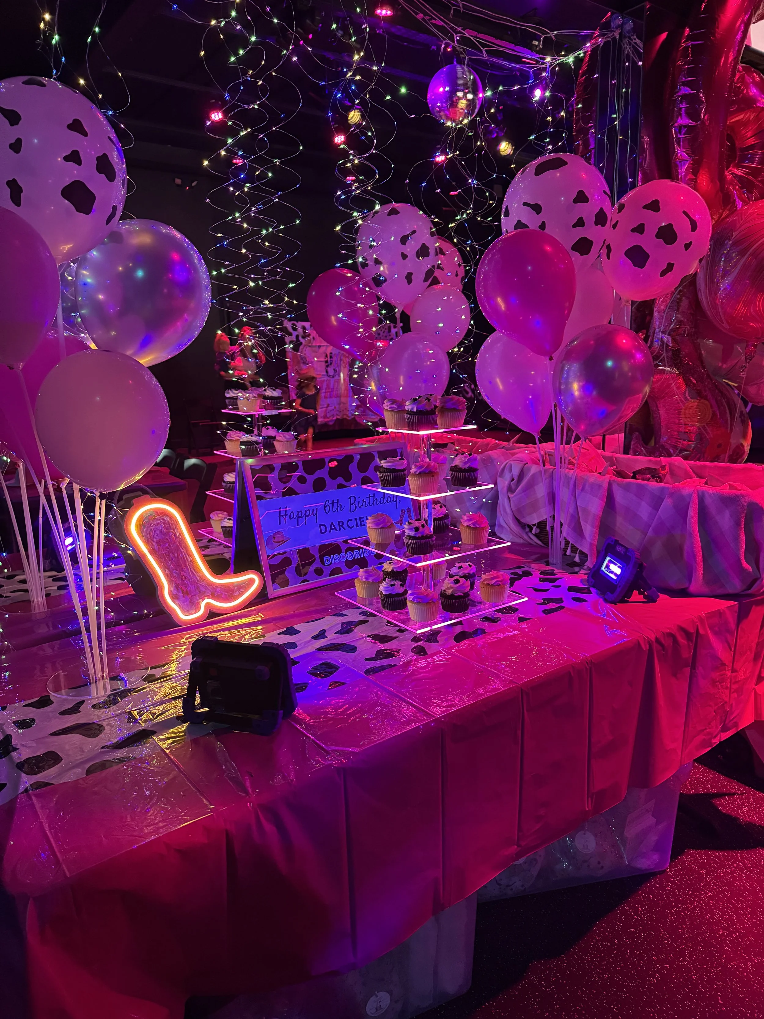 Decorated party table with pink and purple balloons, string lights, and a cake stand, celebrating a 6th birthday with a birthday sign, cupcakes, and neon lights.