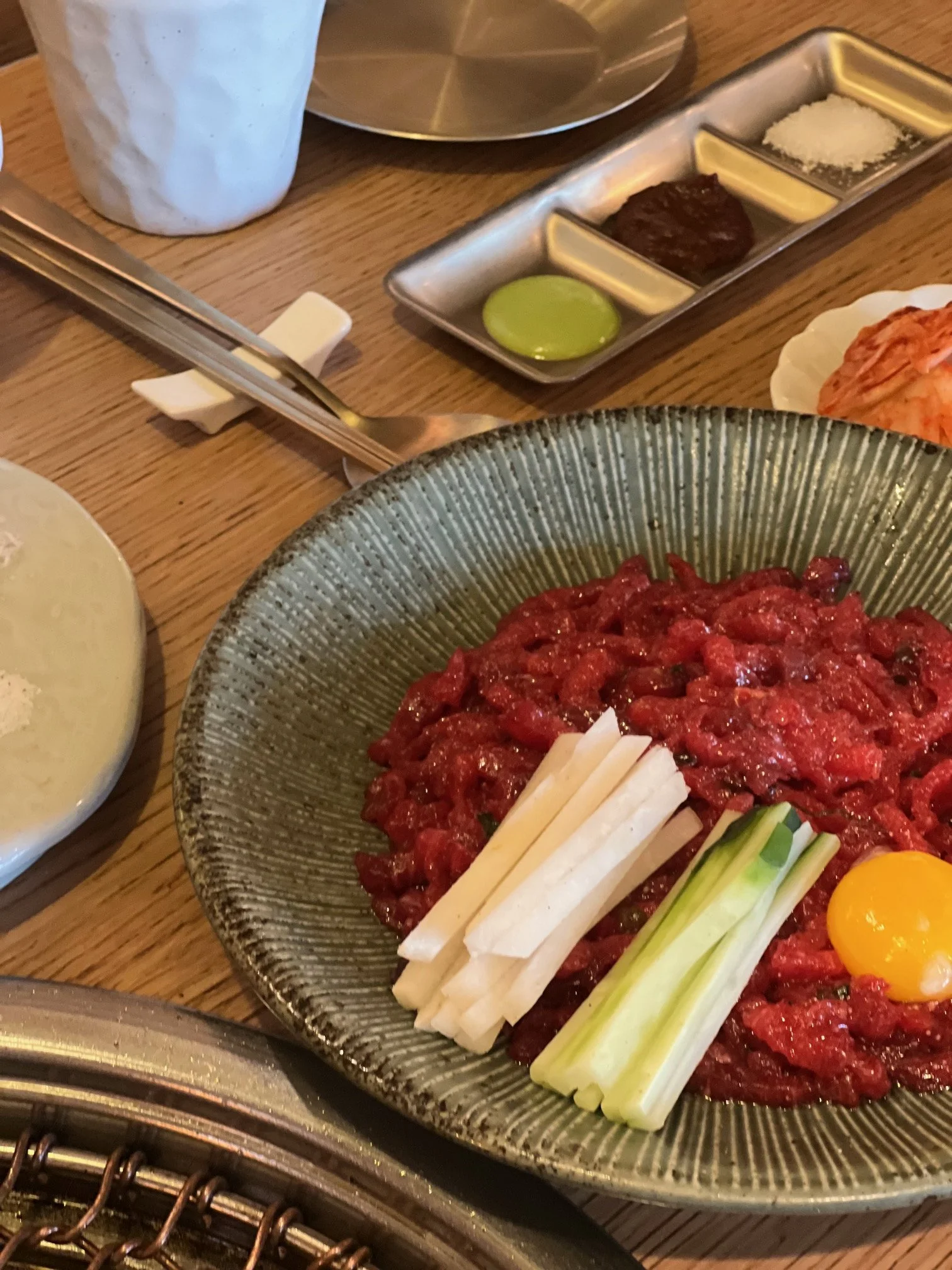 A bowl of raw beef tartare topped with a raw egg yolk, julienned white radish, and sliced celery stalks, with other dishes and condiments on a wooden table.