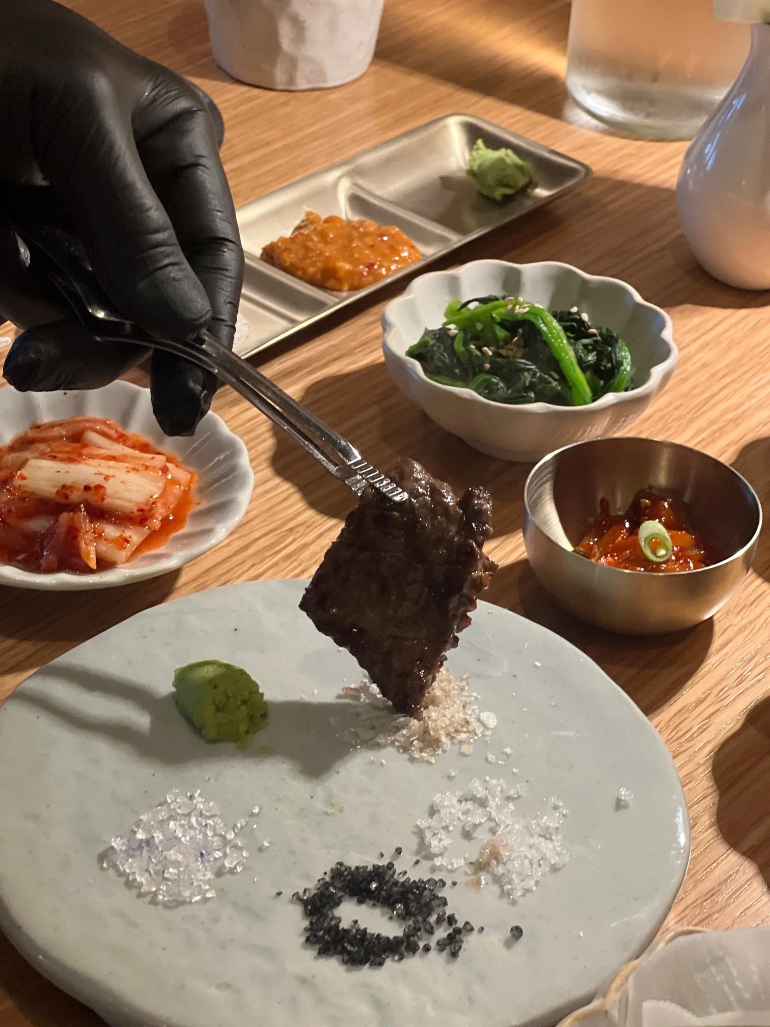 A hand in a black glove uses tongs to pick up a piece of cooked meat from a white plate that has salt, wasabi, and black pepper on it. Various small bowls and dishes with Korean side dishes such as kimchi, spinach, and radish, along with dipping sauc
