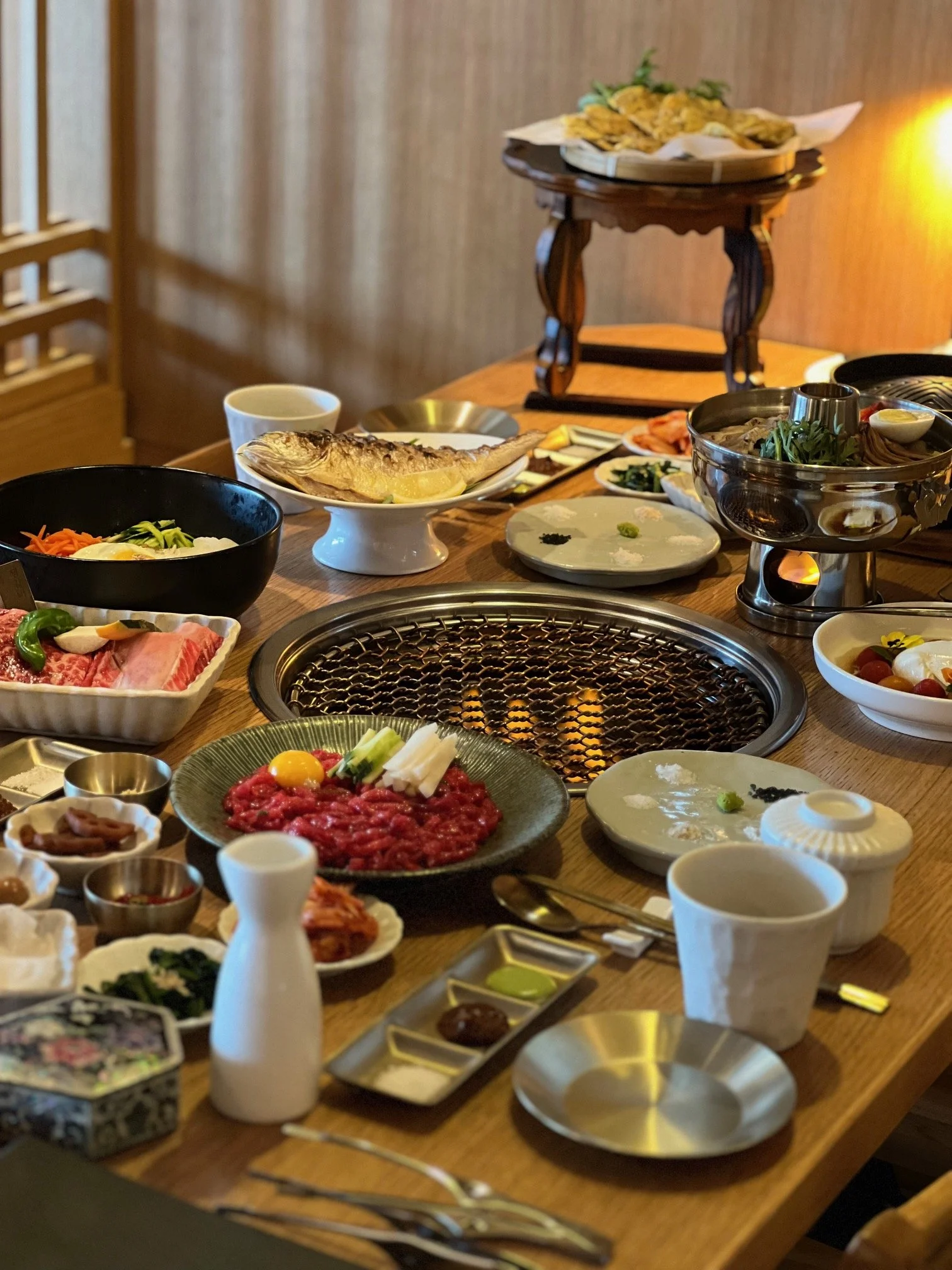 A table set for a Japanese barbecue meal with various raw meats, vegetables, sauces, and a grill in the center.