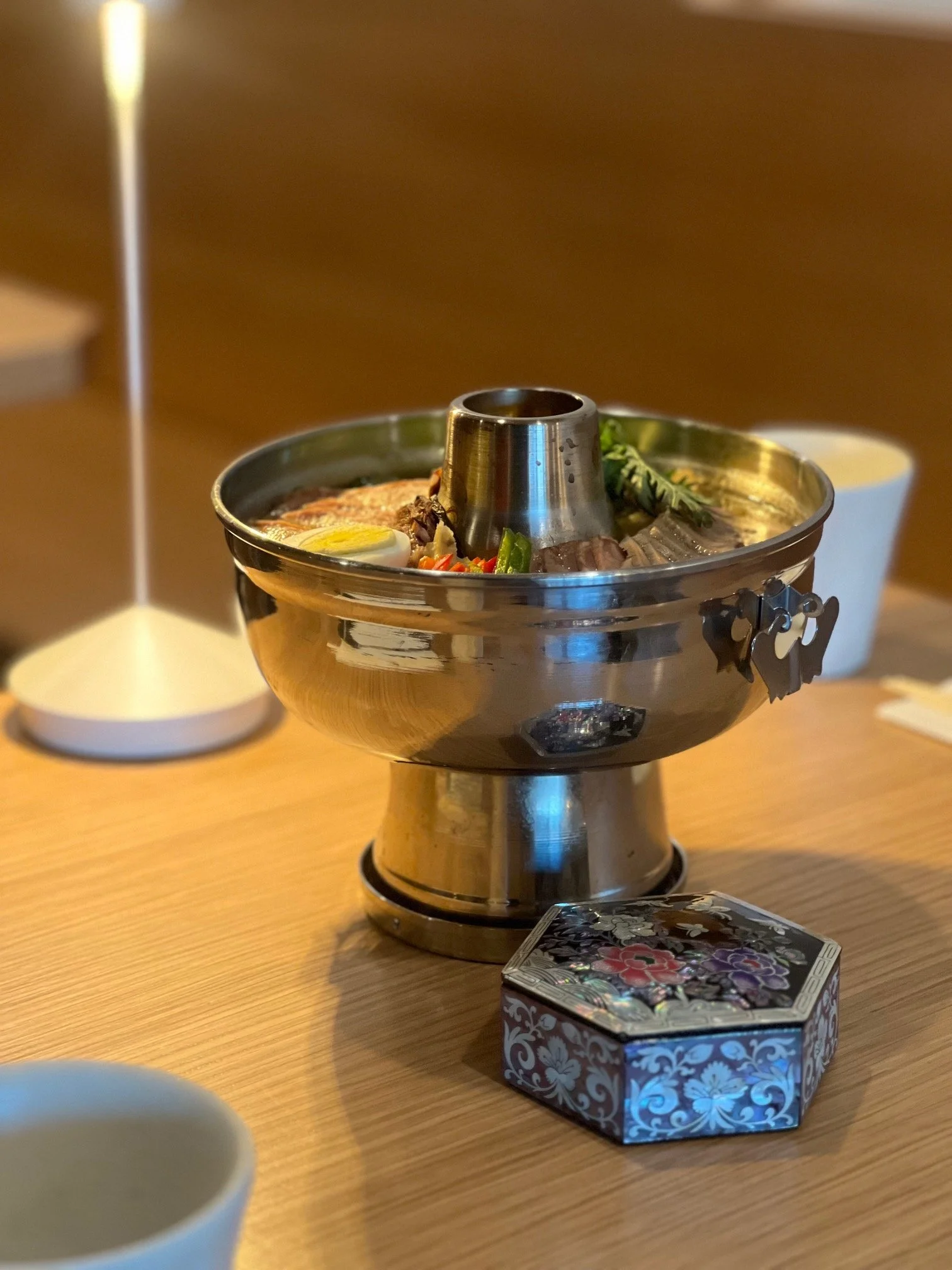 A steaming bowl of hot pot with vegetables and meat, placed on a wooden table with a decorative box nearby.
