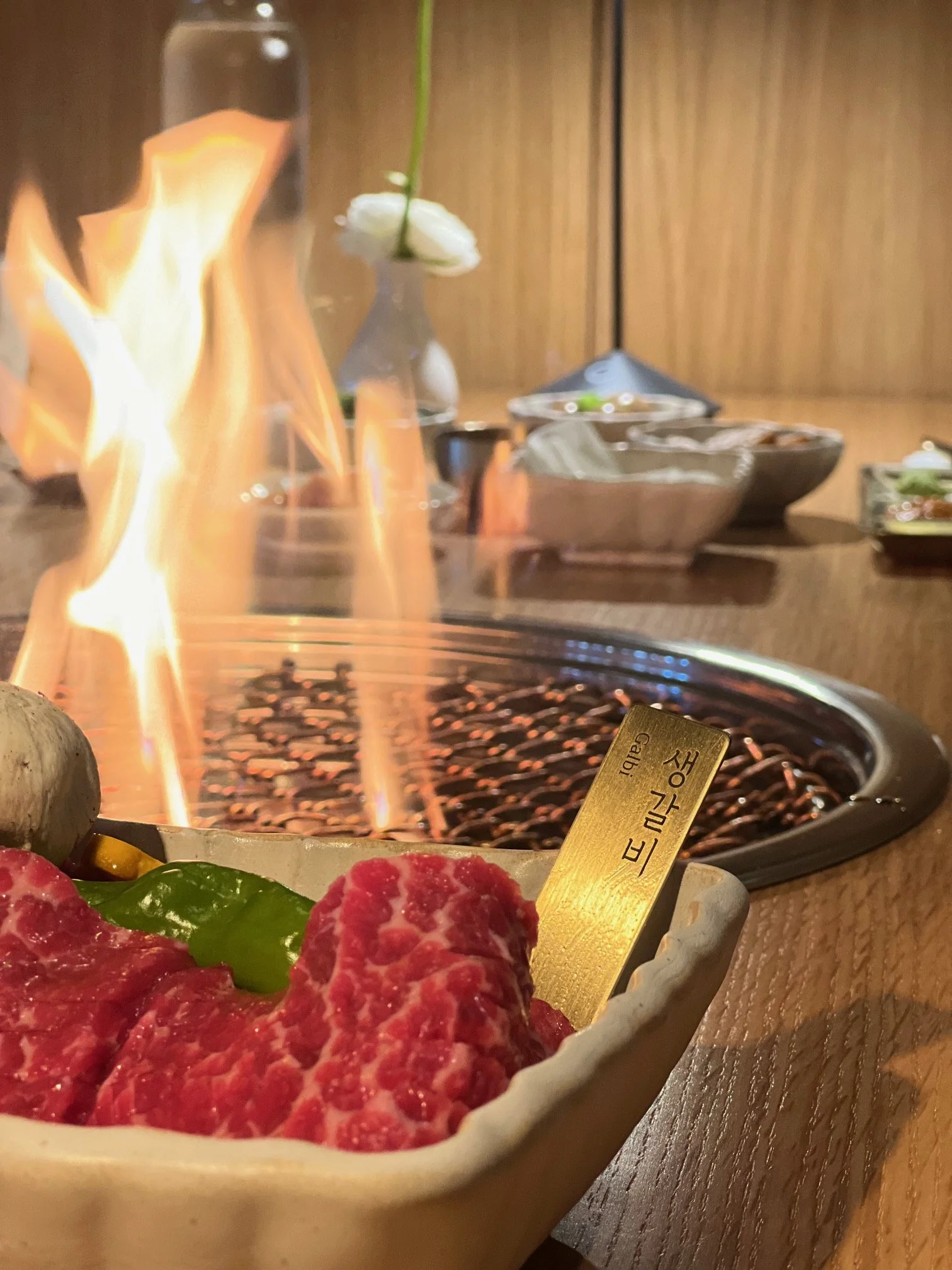 Raw beef and vegetables on a plate near a tabletop grill with flames, in a dining setting.